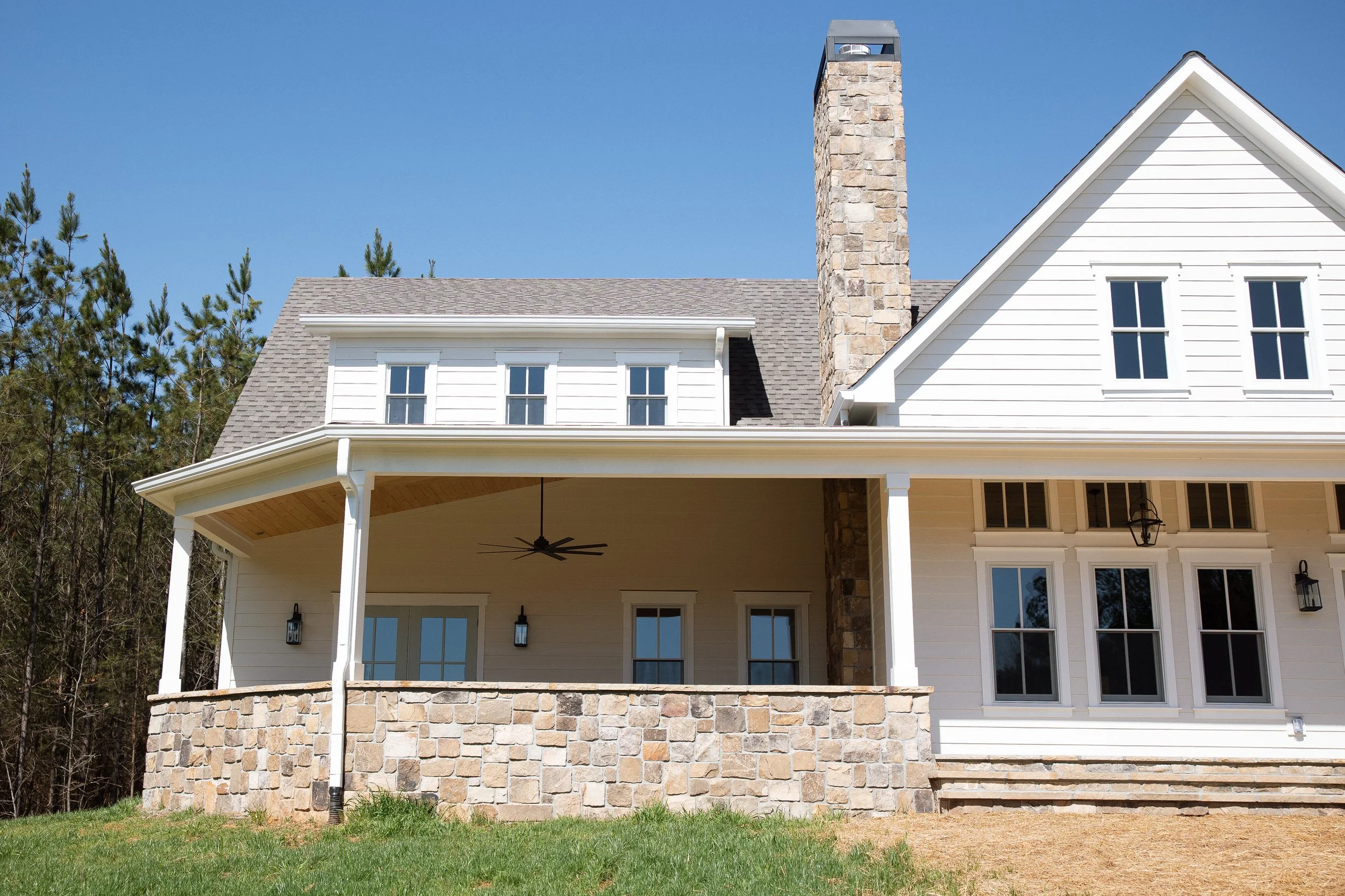 A large two-story house with white siding and gray roof, stone chimney, covered porch with ceiling fan, outdoor light fixtures, and stairs leading to the yard. Background features trees and a clear blue sky.