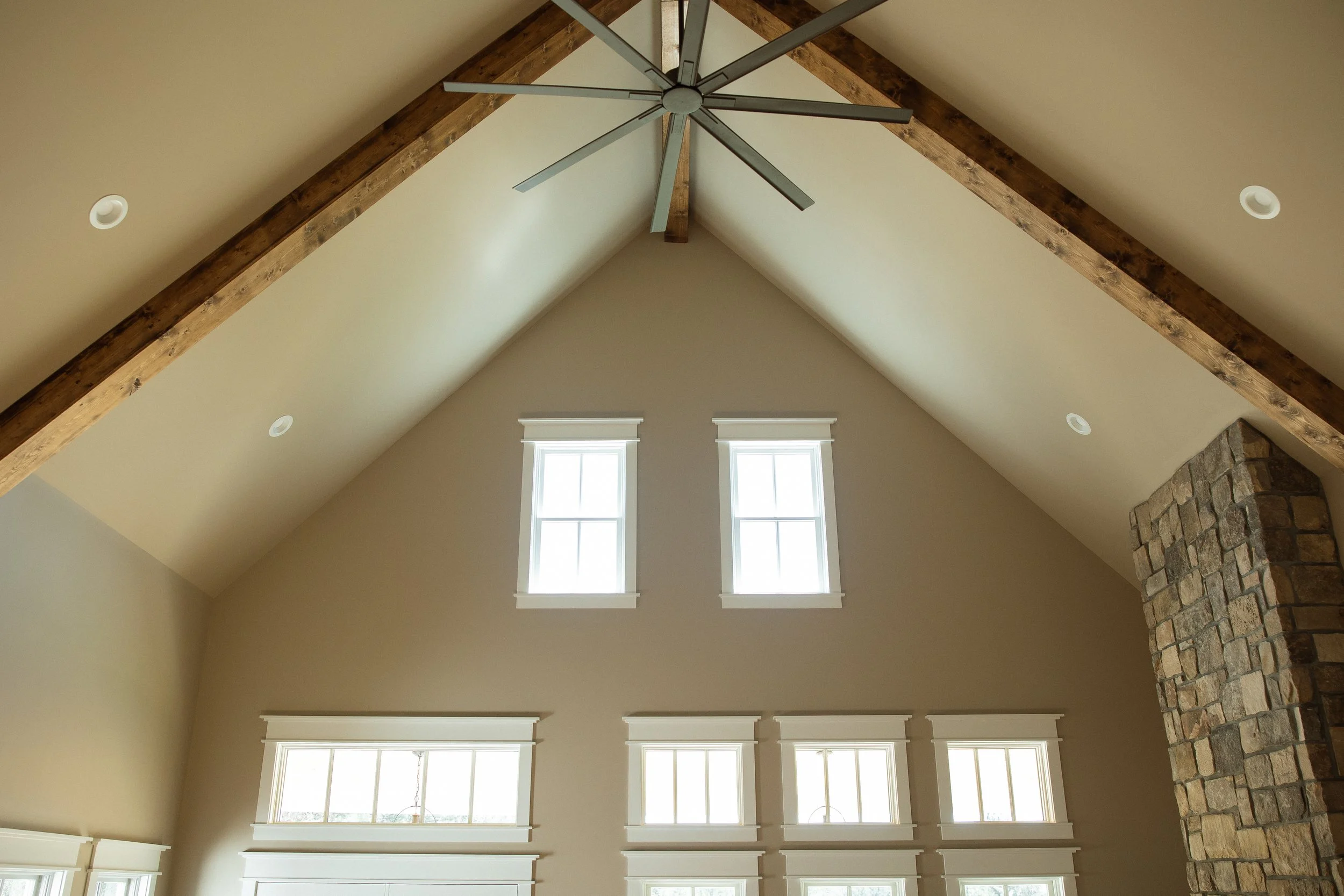 Interior view of a vaulted ceiling with exposed wooden beams, a modern ceiling fan, and multiple windows with white trim and shutters.