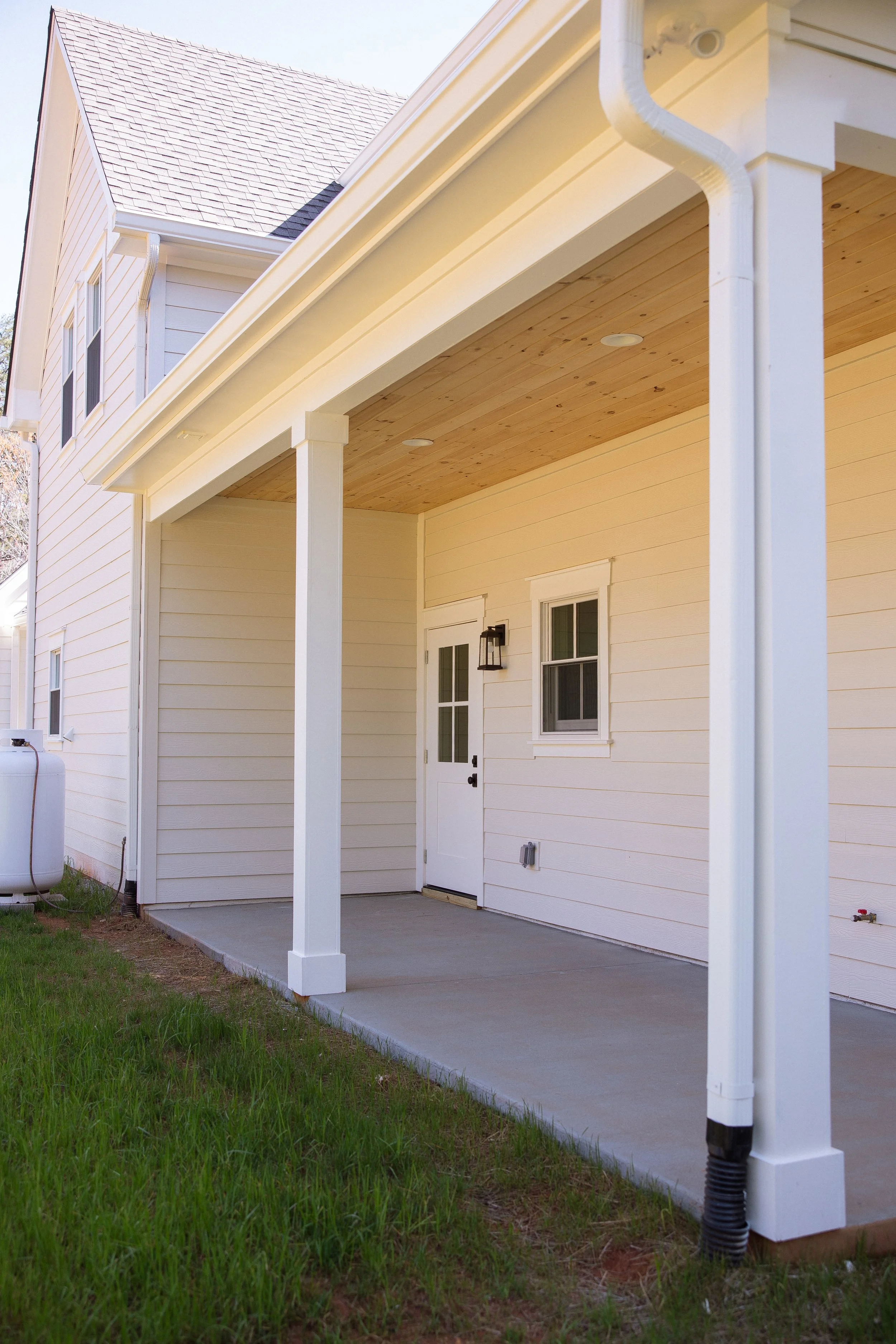 Front porch area of a house with a concrete floor, white siding, a door with a lantern-style light fixture, a small window, and white support columns. Part of a lawn and a propane tank are visible.