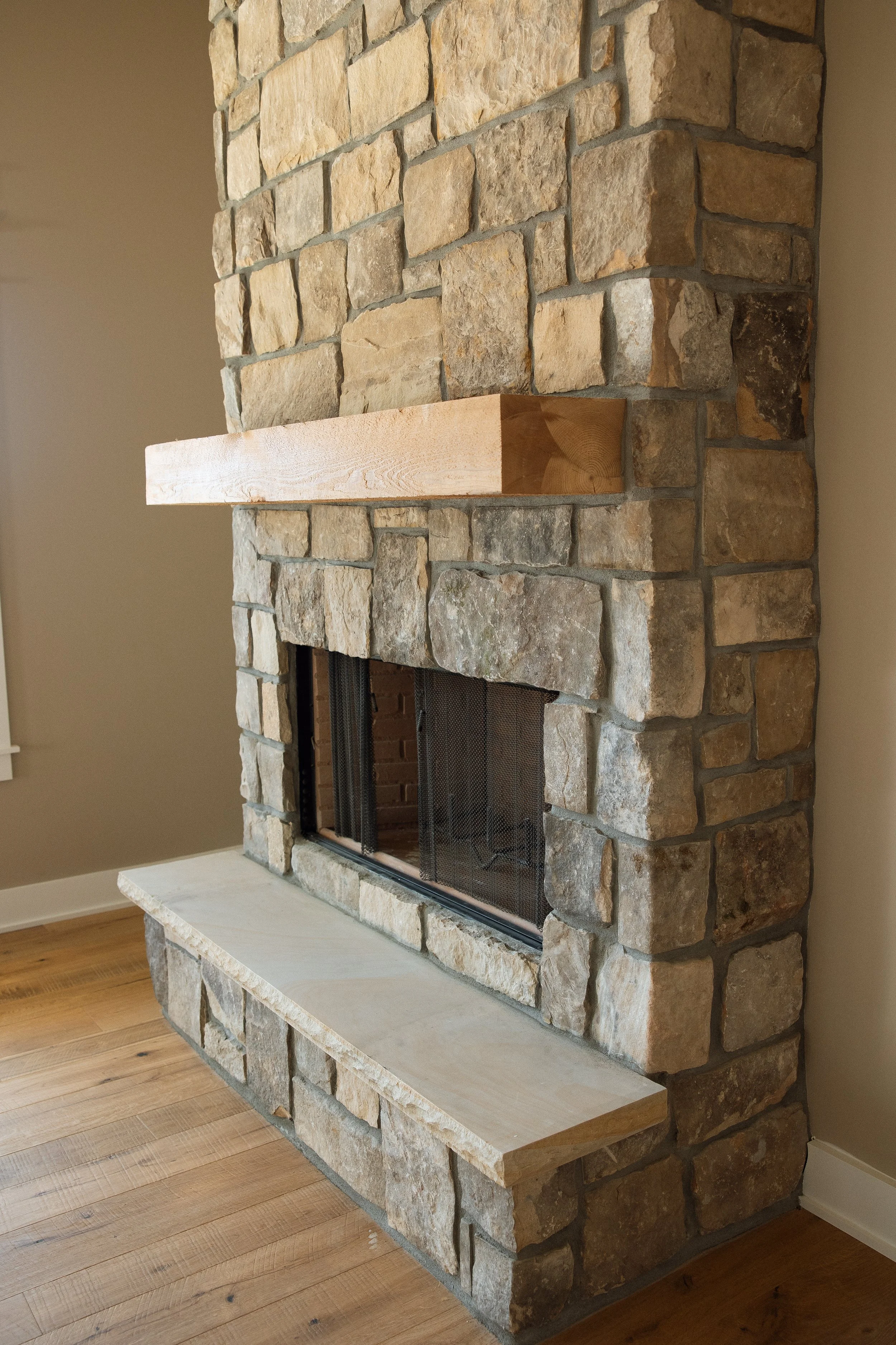 Stone fireplace with wooden mantel and stone hearth, in a room with hardwood floors and beige walls.