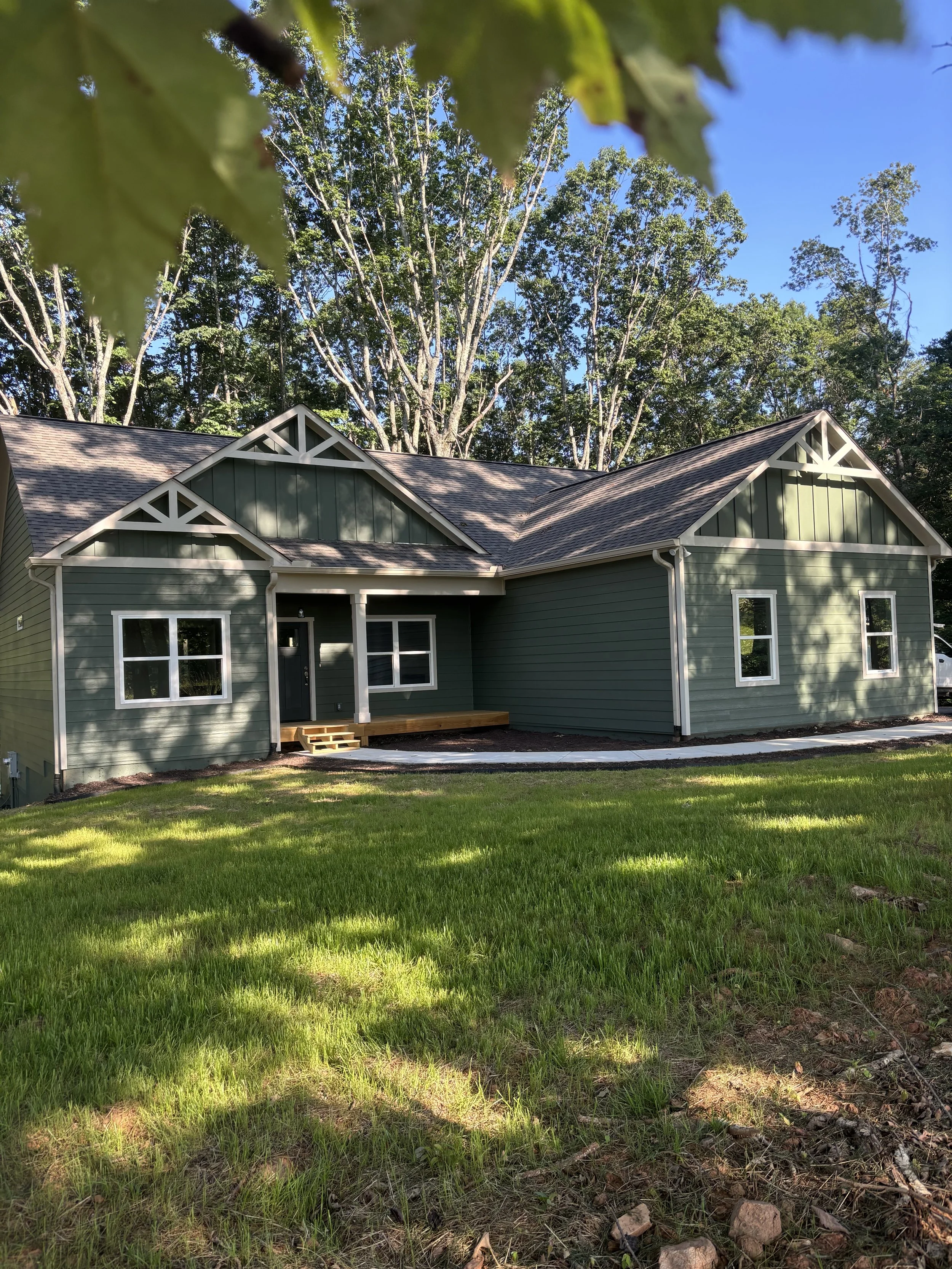 A new green house with white trim and decorative gable details, surrounded by a green lawn and tall trees in the background, under a clear blue sky.
