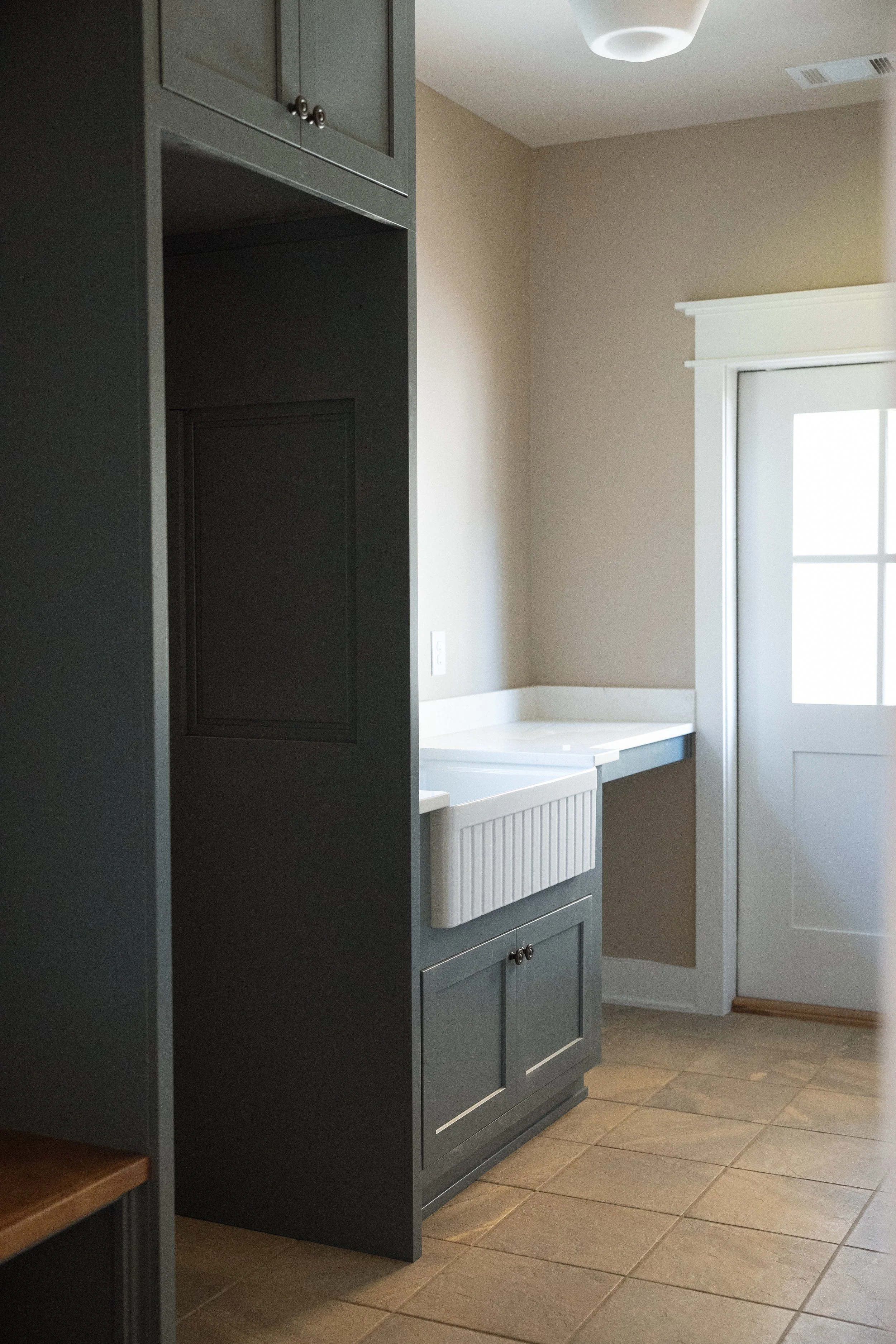 A laundry room with gray cabinets, a white farmhouse sink, beige walls, a white door with window panes, and brown tiled flooring.