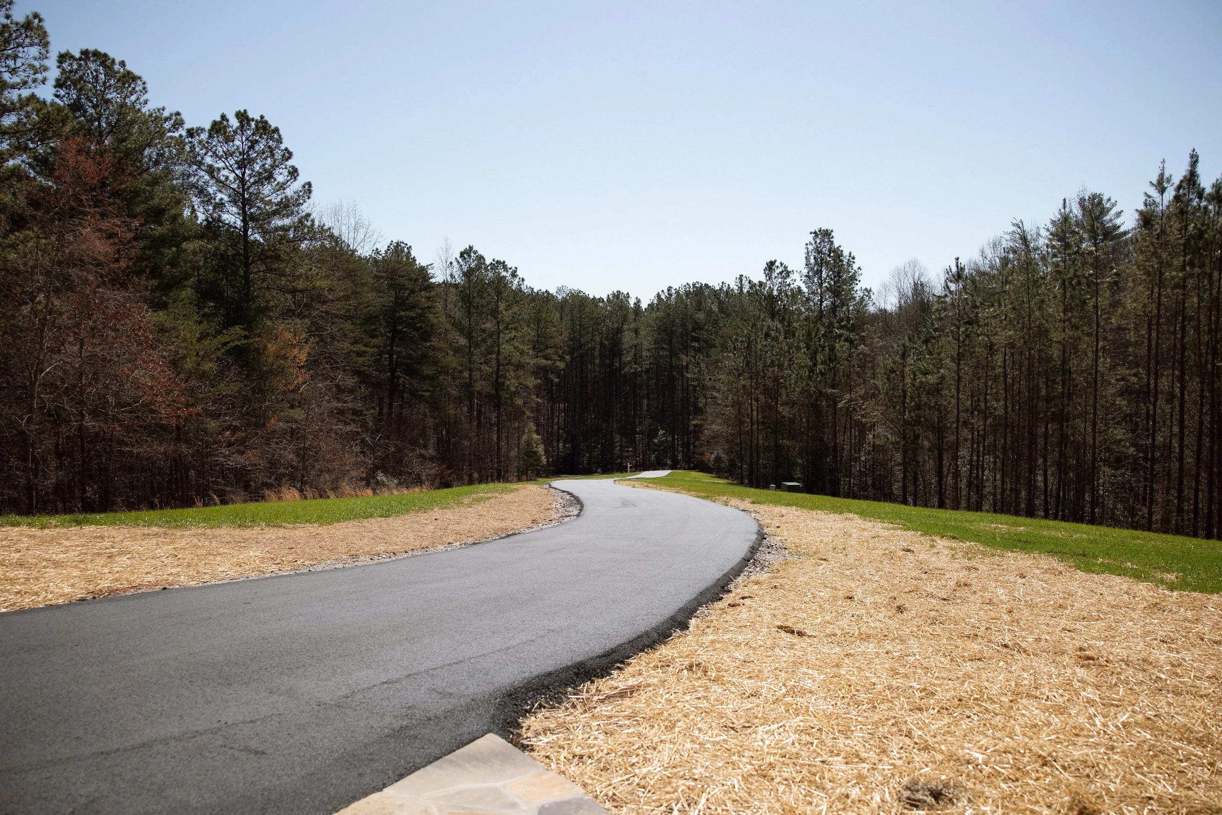 A winding paved road through a wooded landscape with trees on both sides under a clear sky.