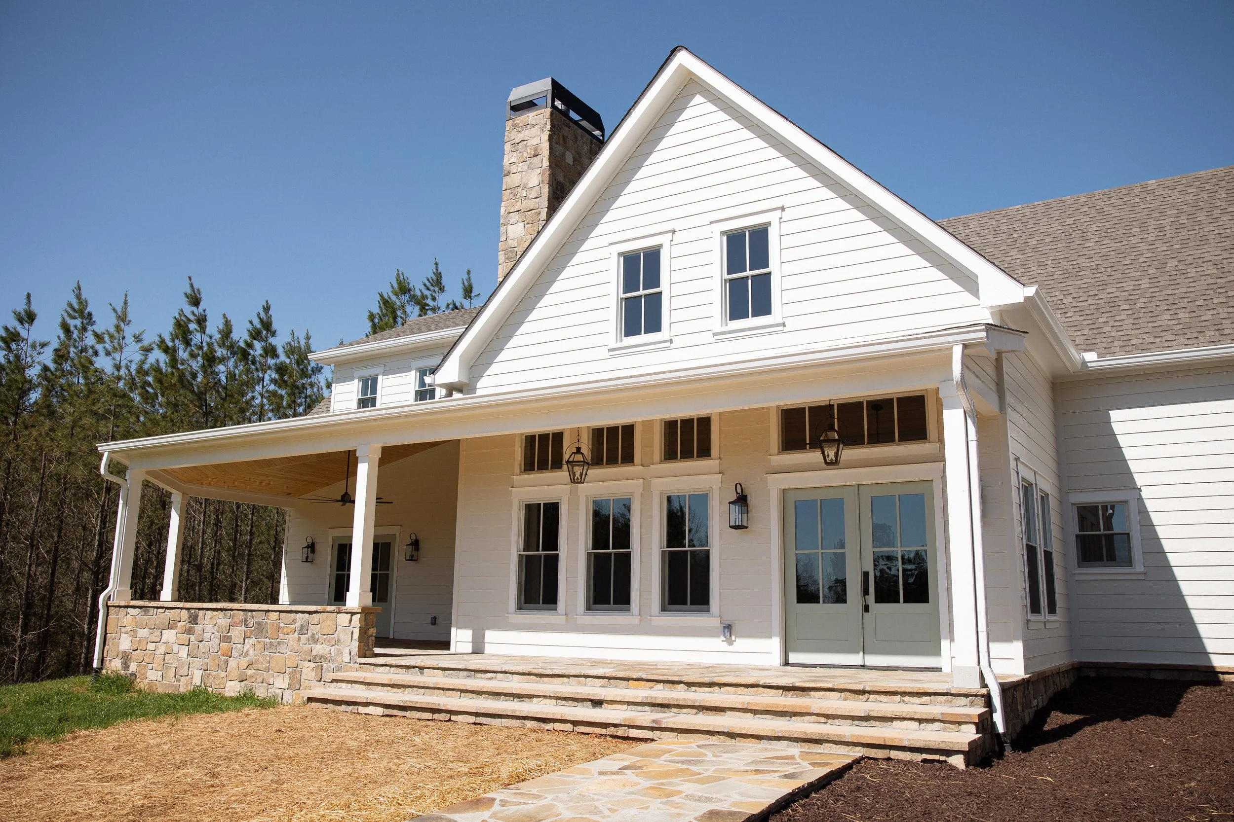 A white two-story house with a gabled roof, multiple windows, and a covered porch with stone steps leading up to it. The porch has hanging lights and a ceiling fan. There are surrounding trees and a clear blue sky.
