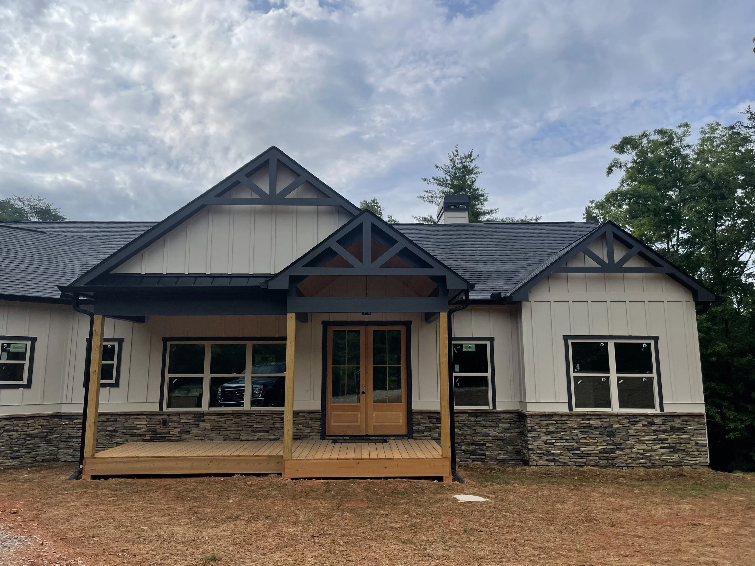 New house under construction with a front porch, stone foundation, and multiple windows, surrounded by trees and an overcast sky.