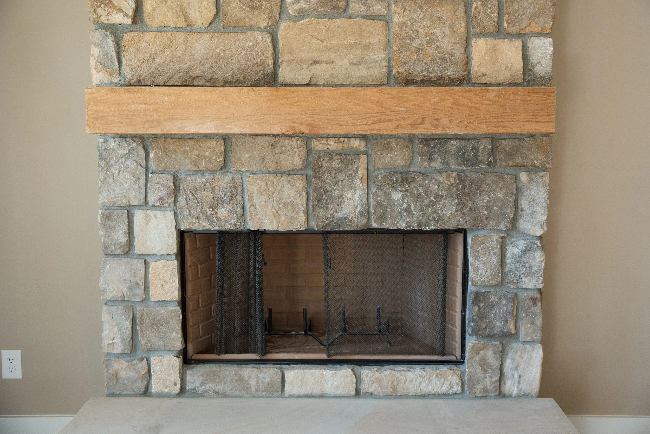 A stone fireplace with a wood mantel in a living room. The firebox has a metal mesh screen and brick interior.