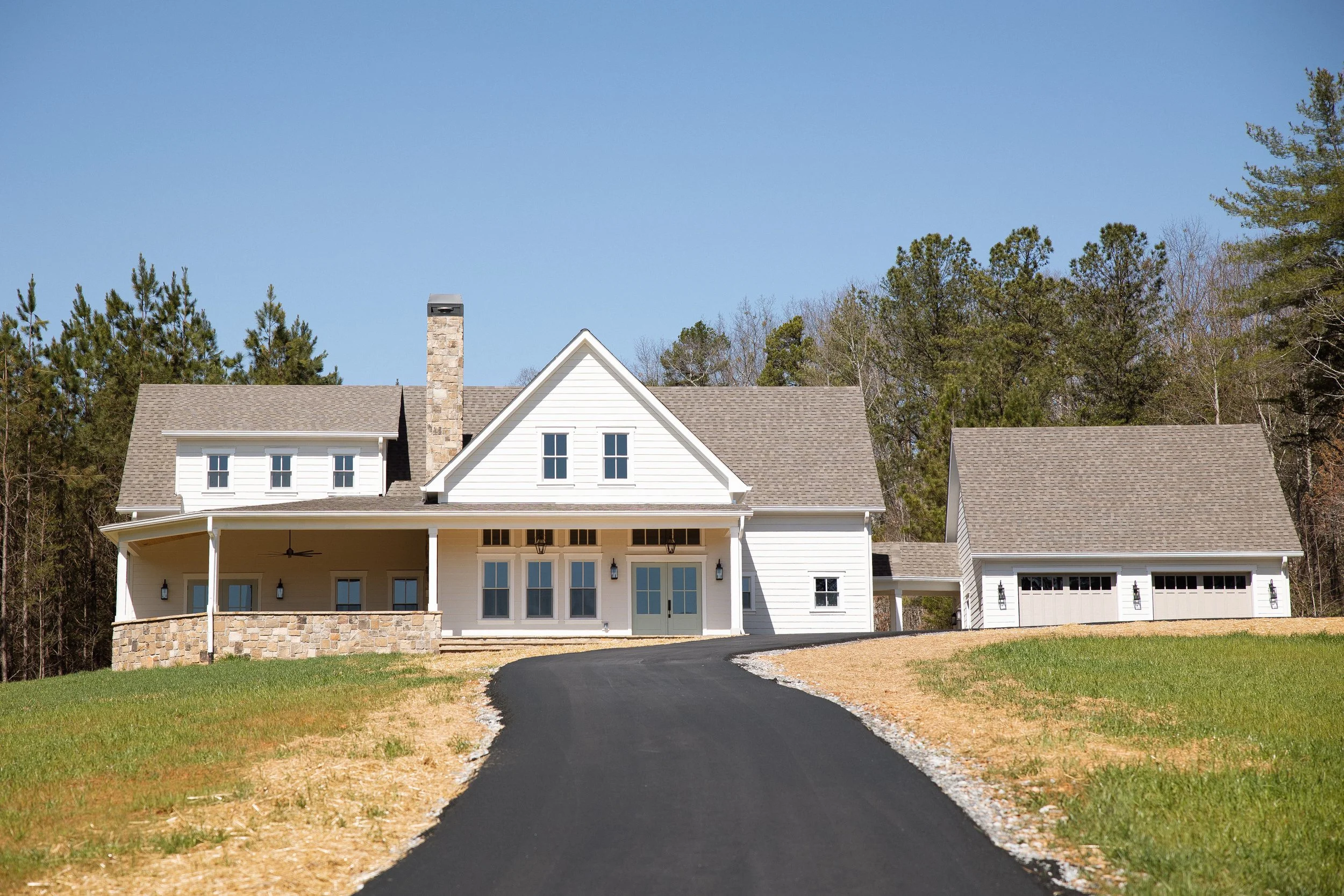 White two-story house with a large porch, chimney, attached garage, and a paved driveway leading up to it, surrounded by a grassy yard and trees in the background.