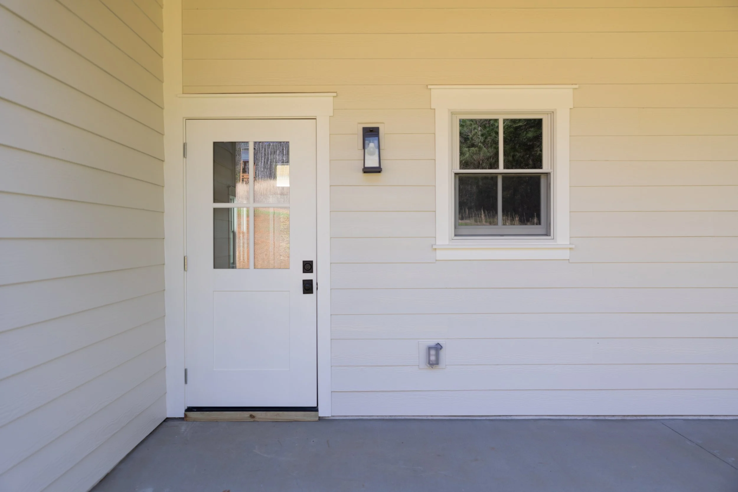 Front porch area of a house featuring a white door with a small window, a white-framed window with four panes, a black outdoor wall light, and a small outdoor wall-mounted light fixture, with a concrete floor.