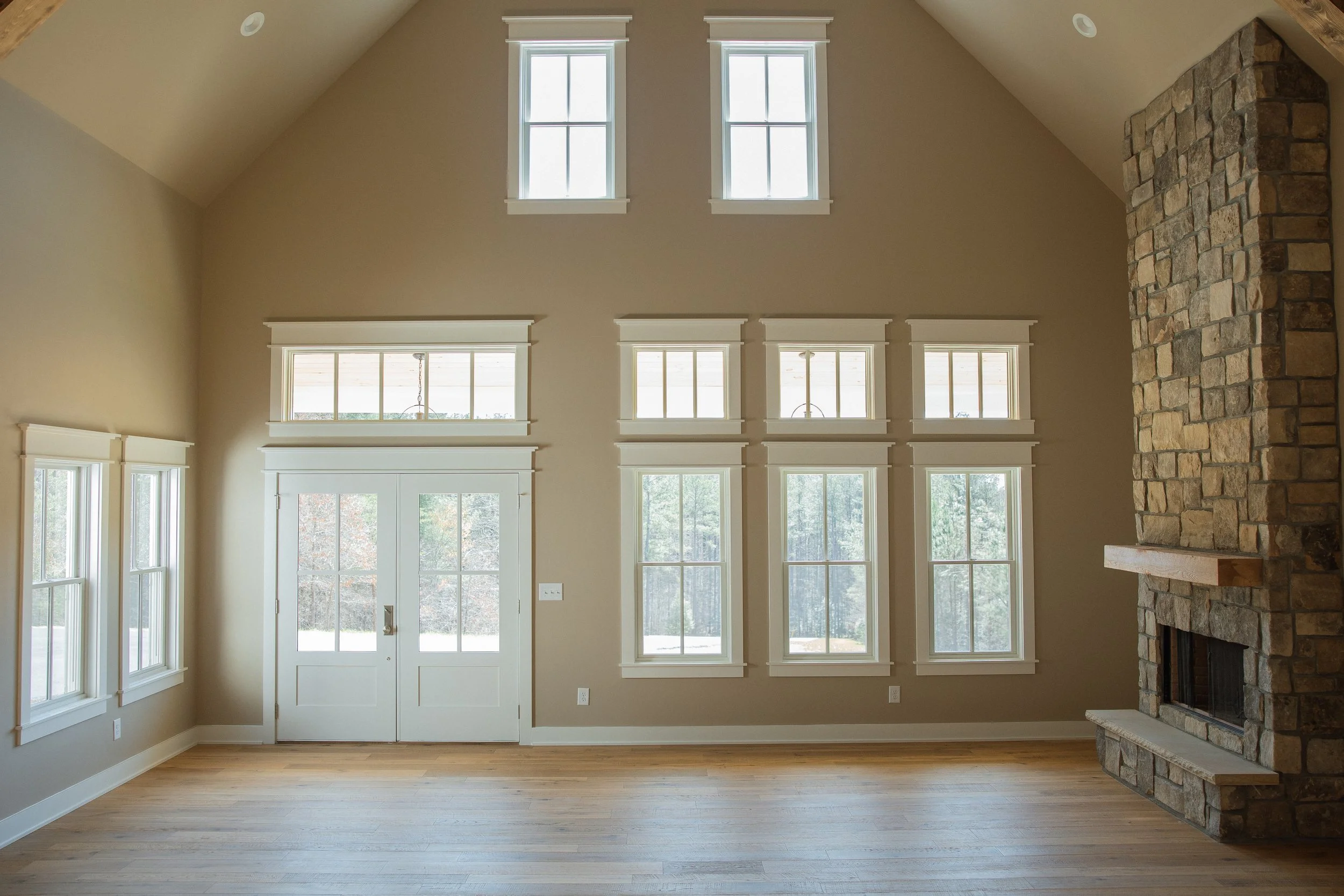 Empty living room with a high ceiling, beige walls, multiple large windows, and a stone fireplace.