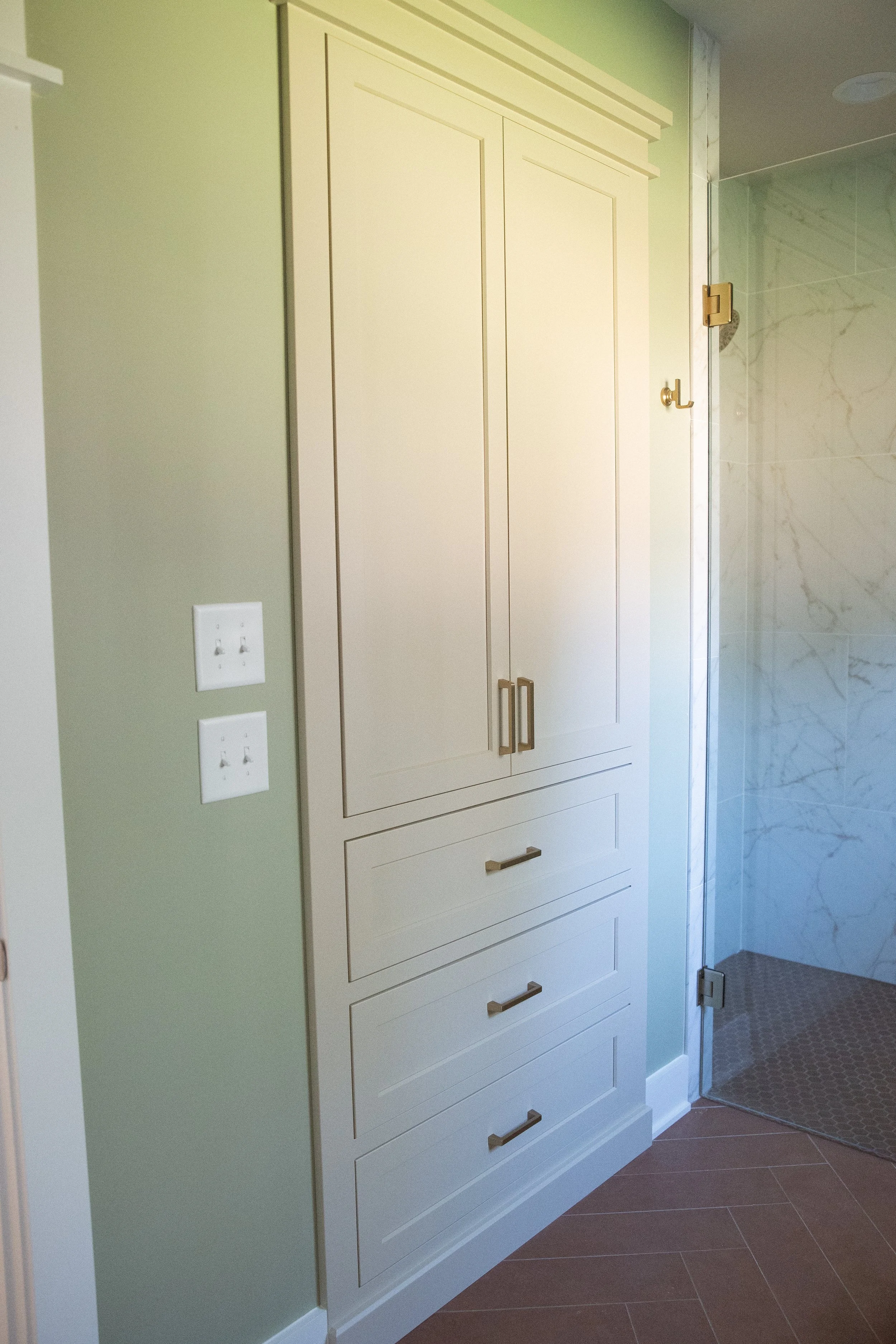 Bathroom storage cabinet with multiple drawers and a door, near a glass-enclosed shower with marble walls.