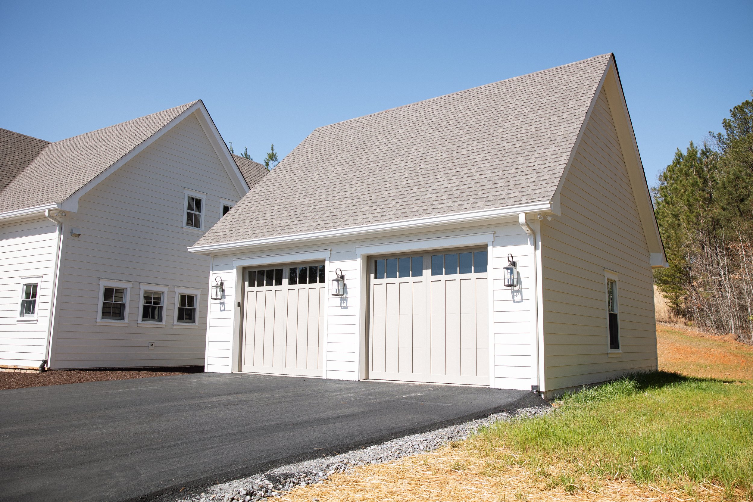 White garage with two doors and outdoor lamps, paved driveway, grass and trees in the background under a clear blue sky.