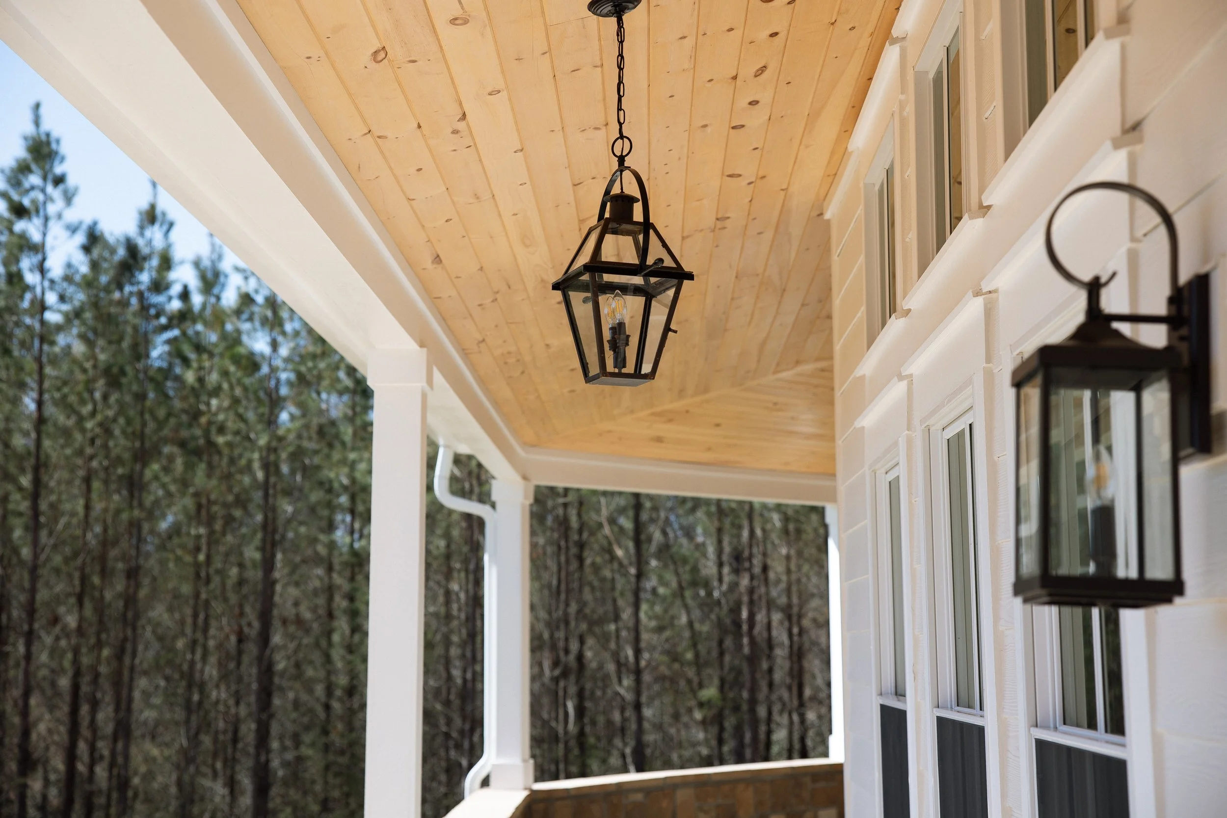 Close-up of a house porch with black lantern-style outdoor lights, white siding, and a wooden ceiling, with forest trees in the background.