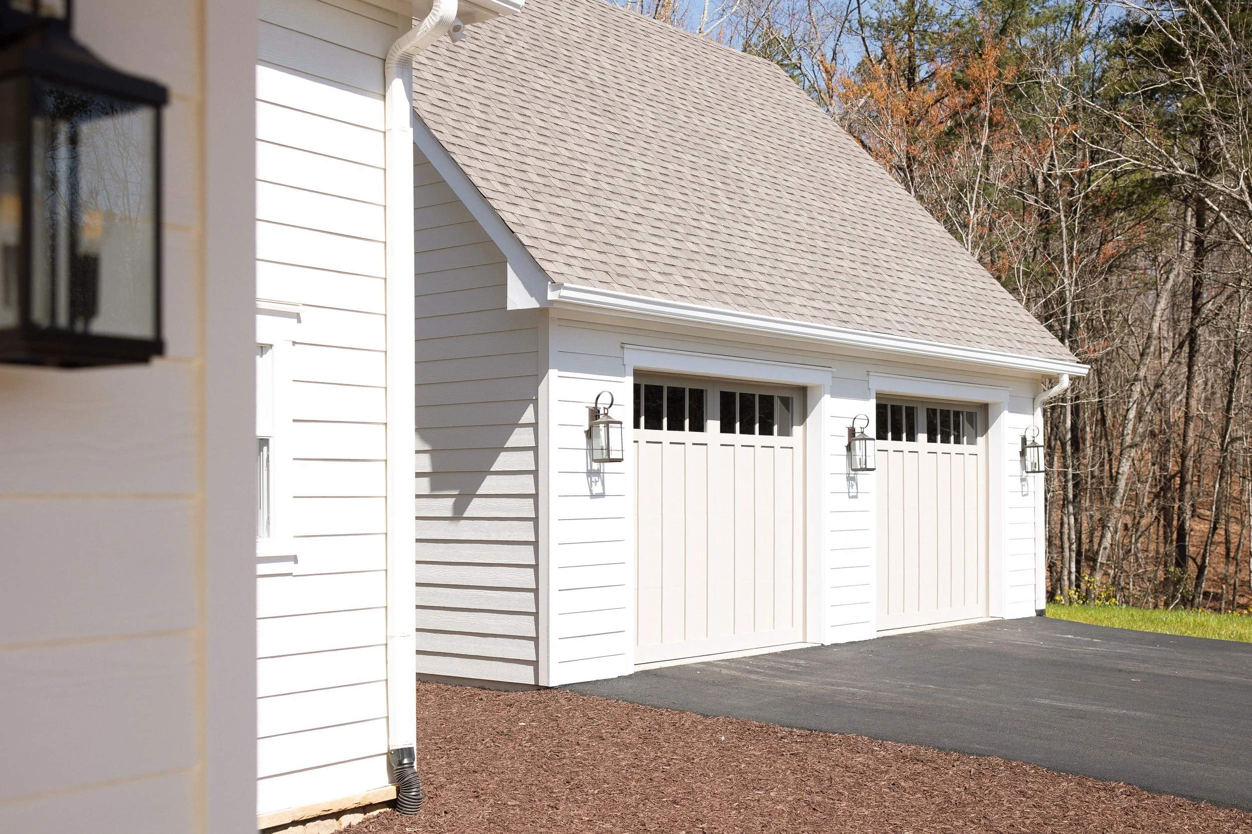 White garage with two doors, exterior lighting, and a paved driveway, with trees in the background.