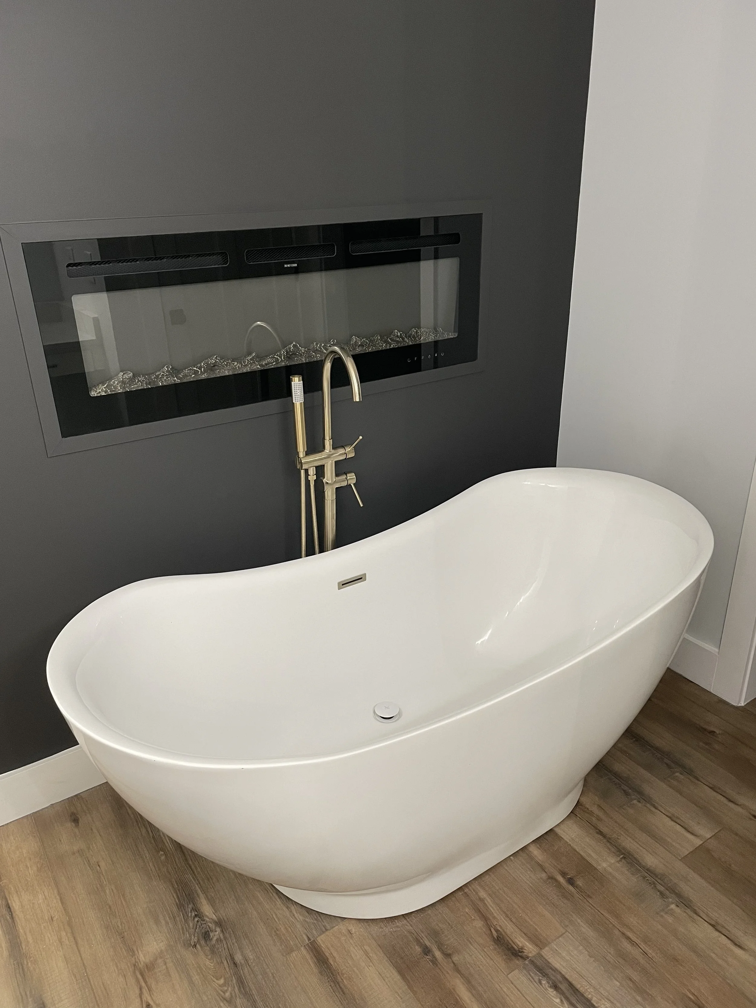 A modern bathroom with a white oval bathtub, a dark gray wall behind it, and a wall-mounted fireplace with glass and stones above the tub. There is a gold-colored faucet with a handheld showerhead attached.