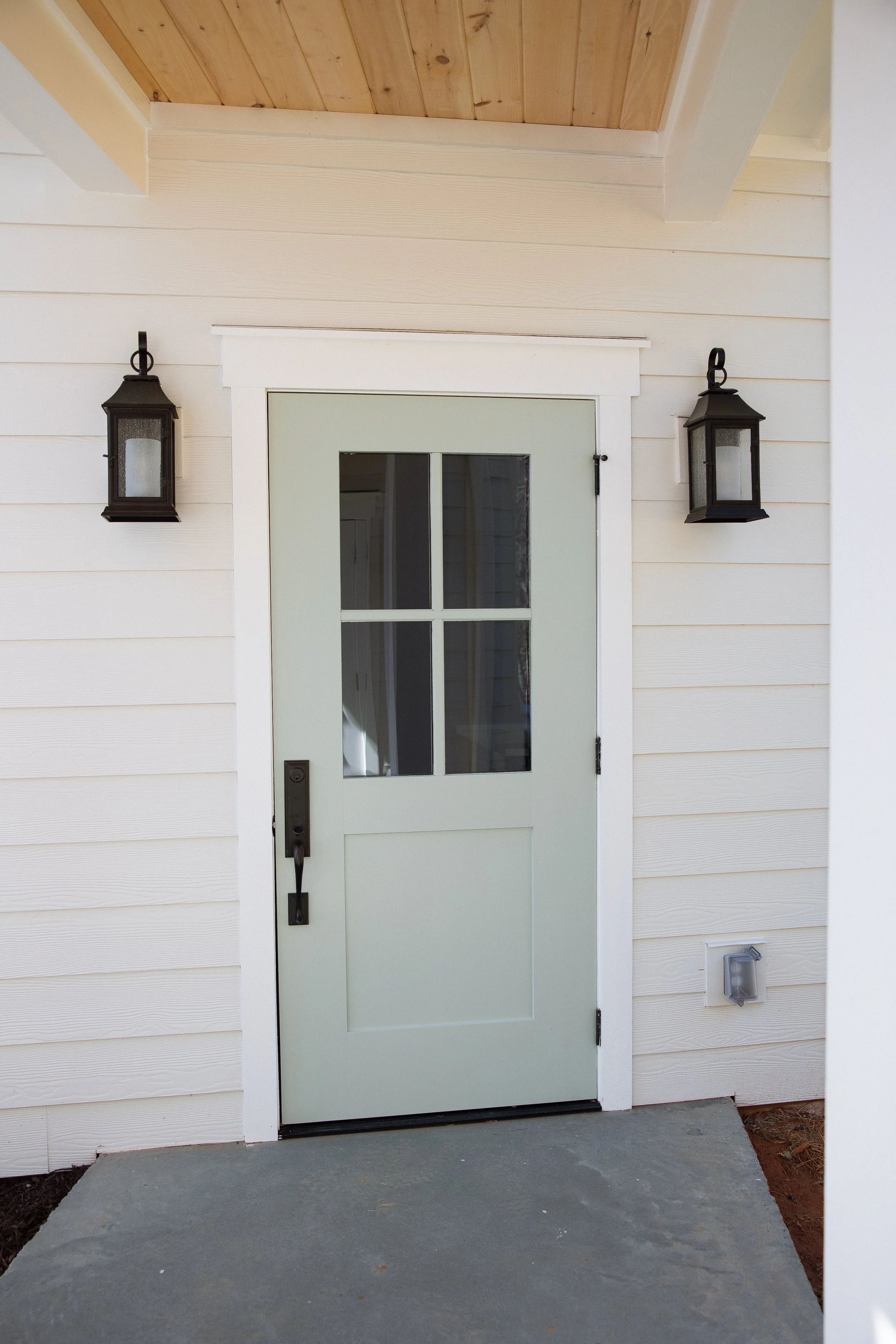 A front door with a glass window set in white siding, flanked by two black lantern-style wall lights, with a concrete porch and a small white electrical box on the wall.