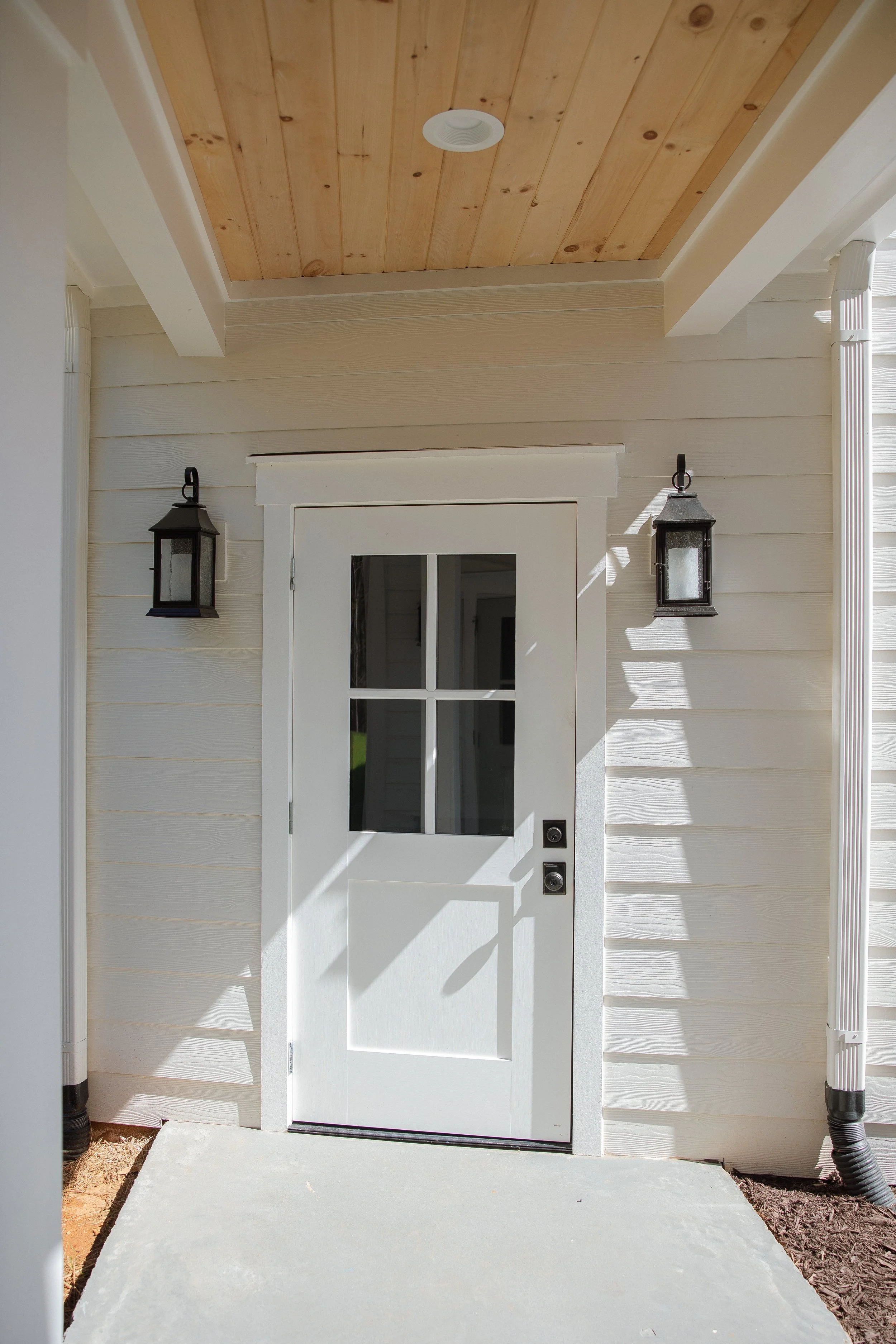 White front door with a small window, flanked by two black outdoor wall lanterns, under a wooden ceiling porch.