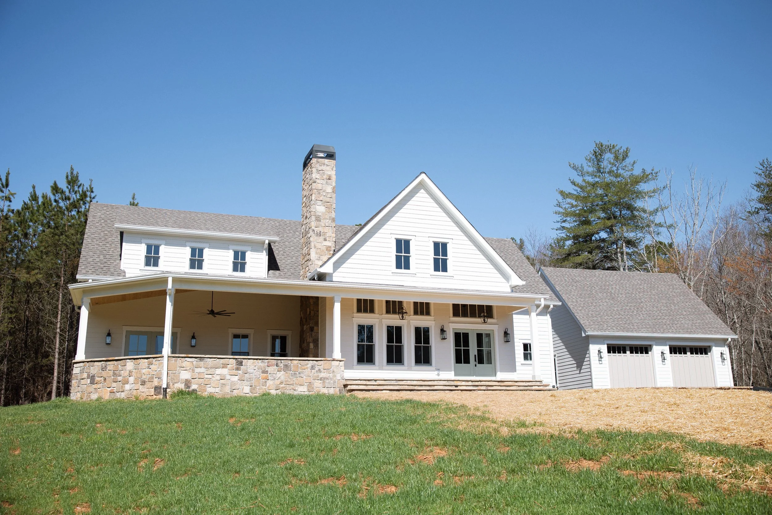A modern white house with a large covered porch, stone chimney, and attached garage, set against a clear blue sky with trees in the background.