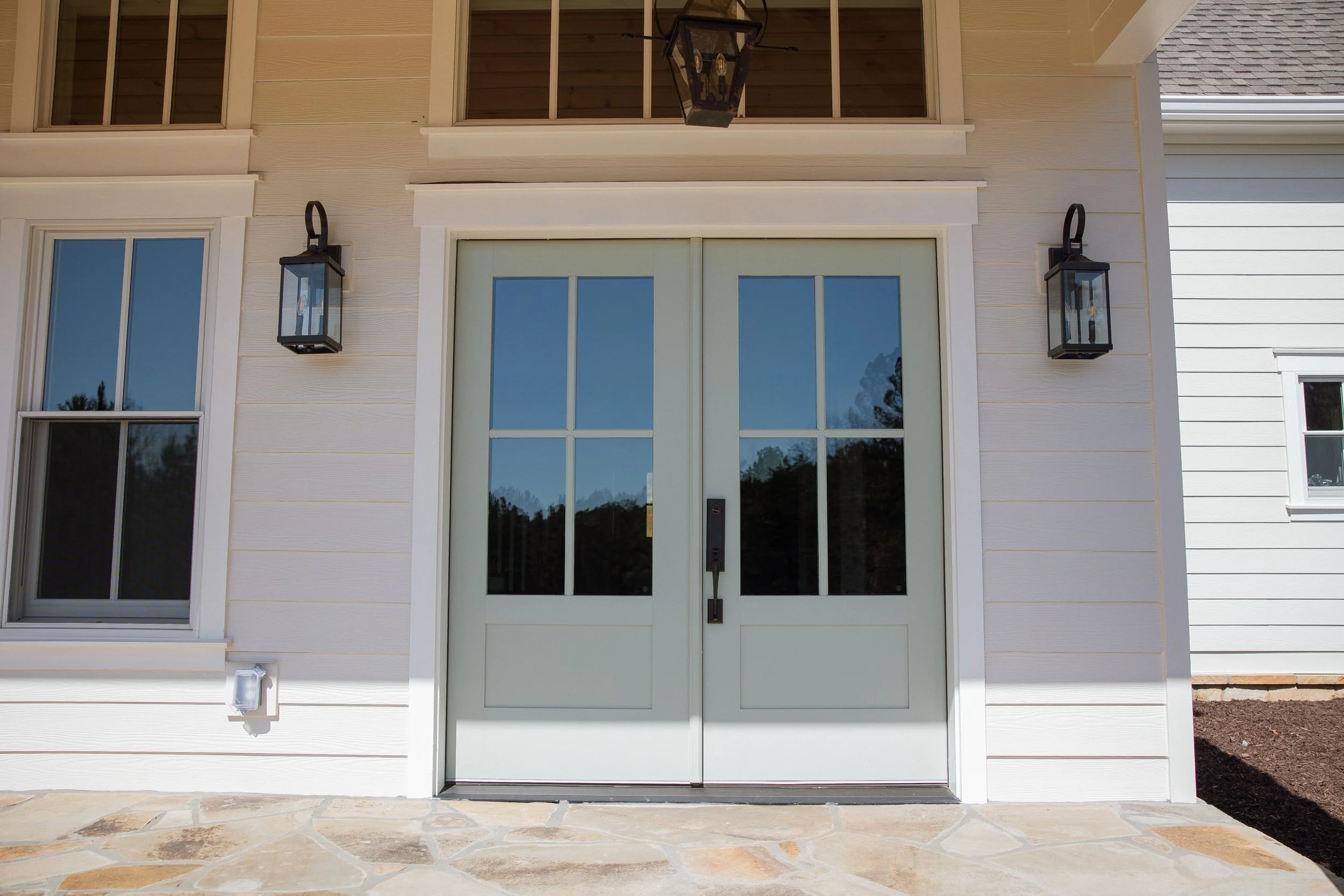 White double front door with large glass panels, flanked by two black outdoor lantern-style wall lights, on a house with white siding and stone pathway.