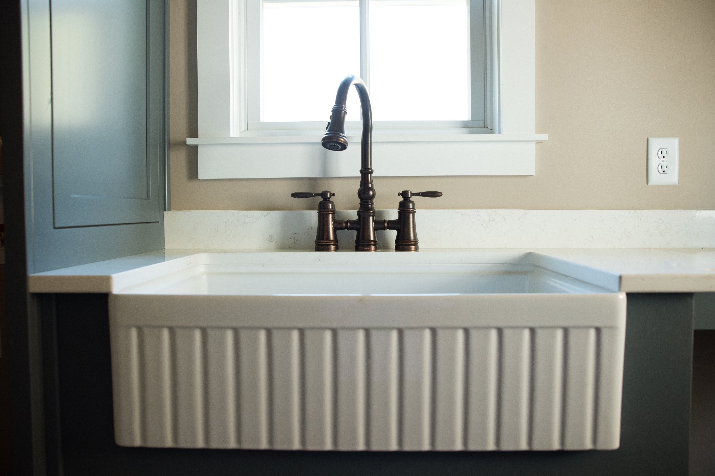 A farmhouse-style white kitchen sink with vertical ridges, a bronze faucet, and a window above it, with a beige wall and an electrical outlet to the right.
