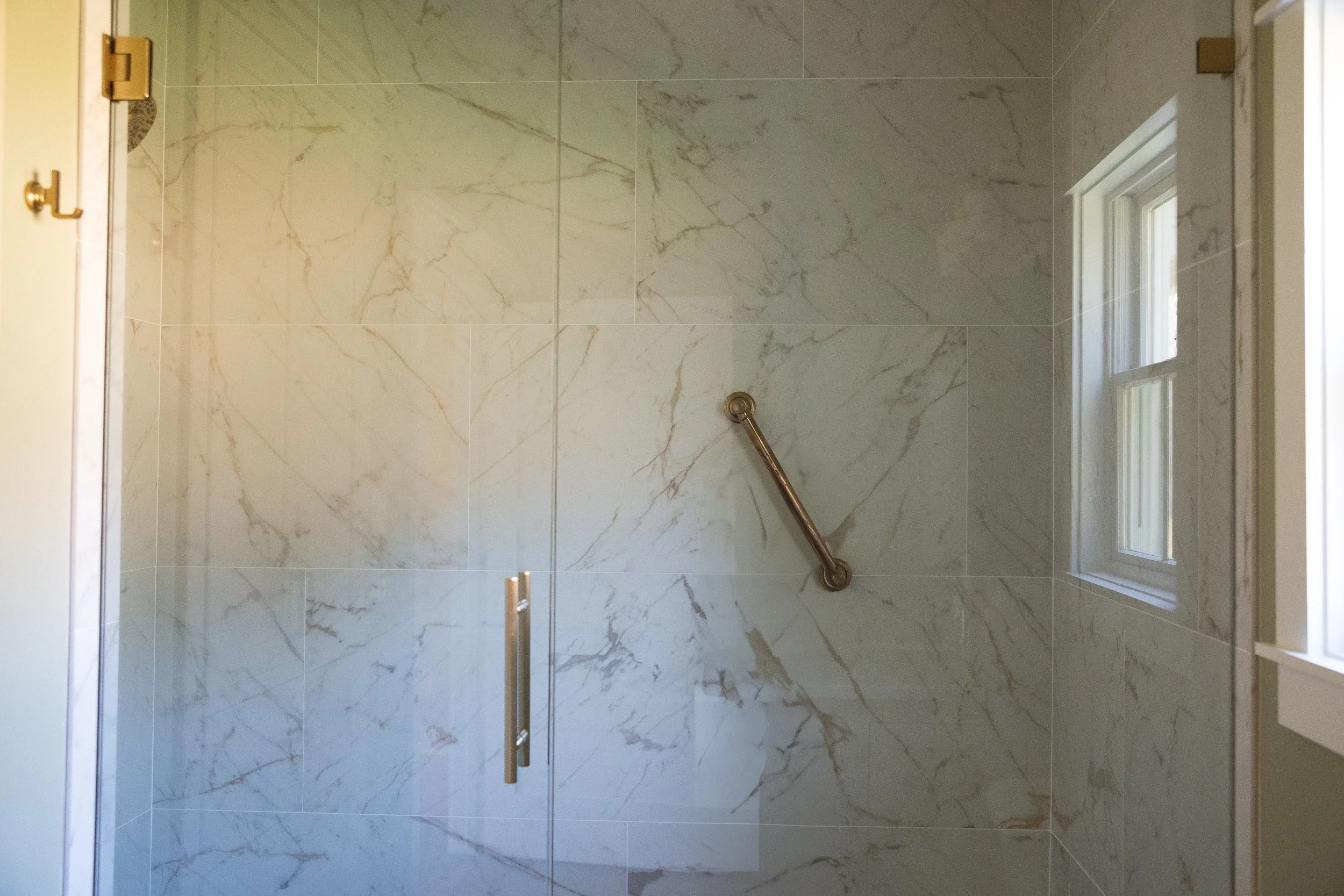 Empty walk-in shower with marble-like tile walls, a metal grab bar, a glass door handle, and a small window.