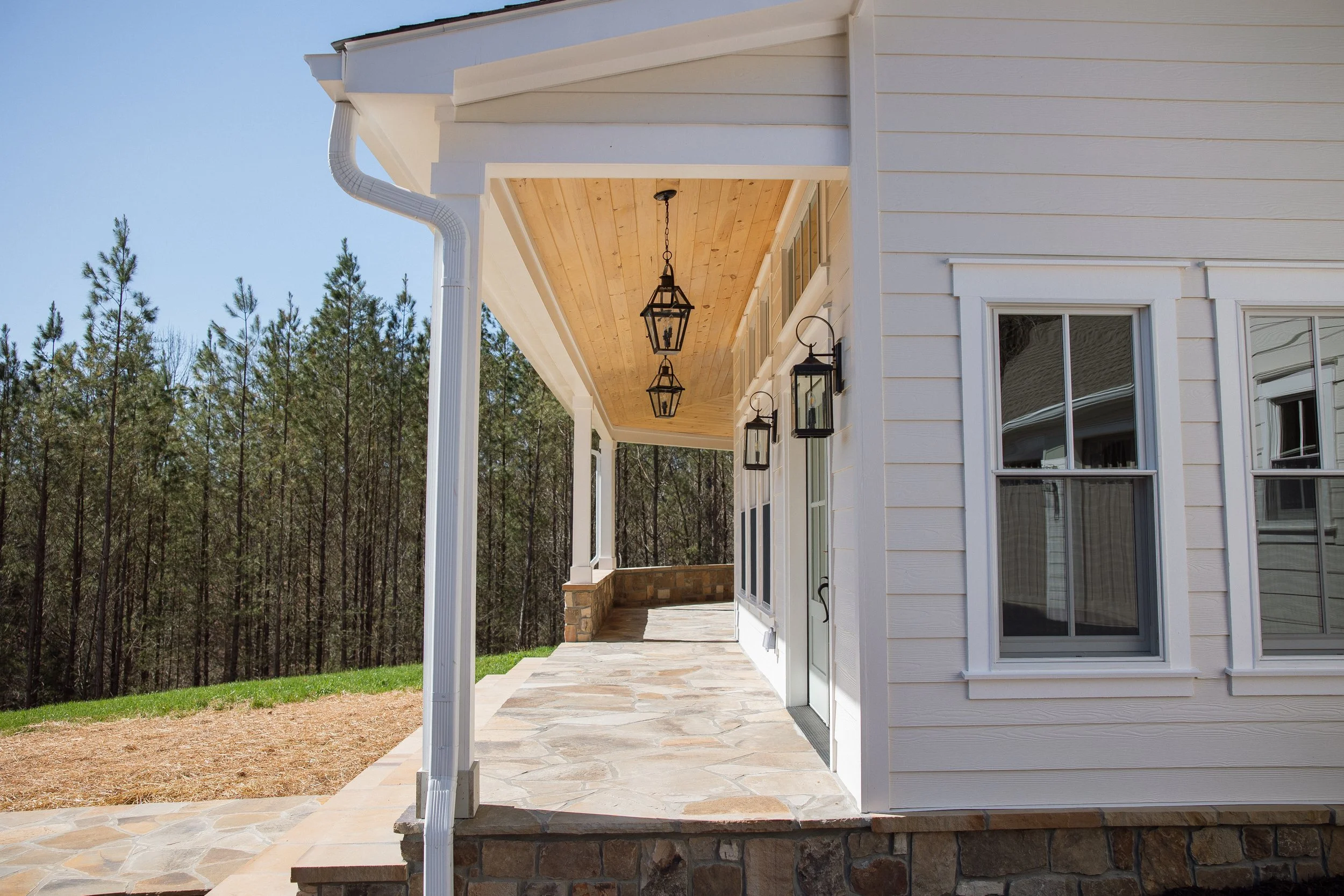 Front porch of a white house with stone patio, hanging lanterns, and wall-mounted lanterns, overlooking a wooded area with tall trees.