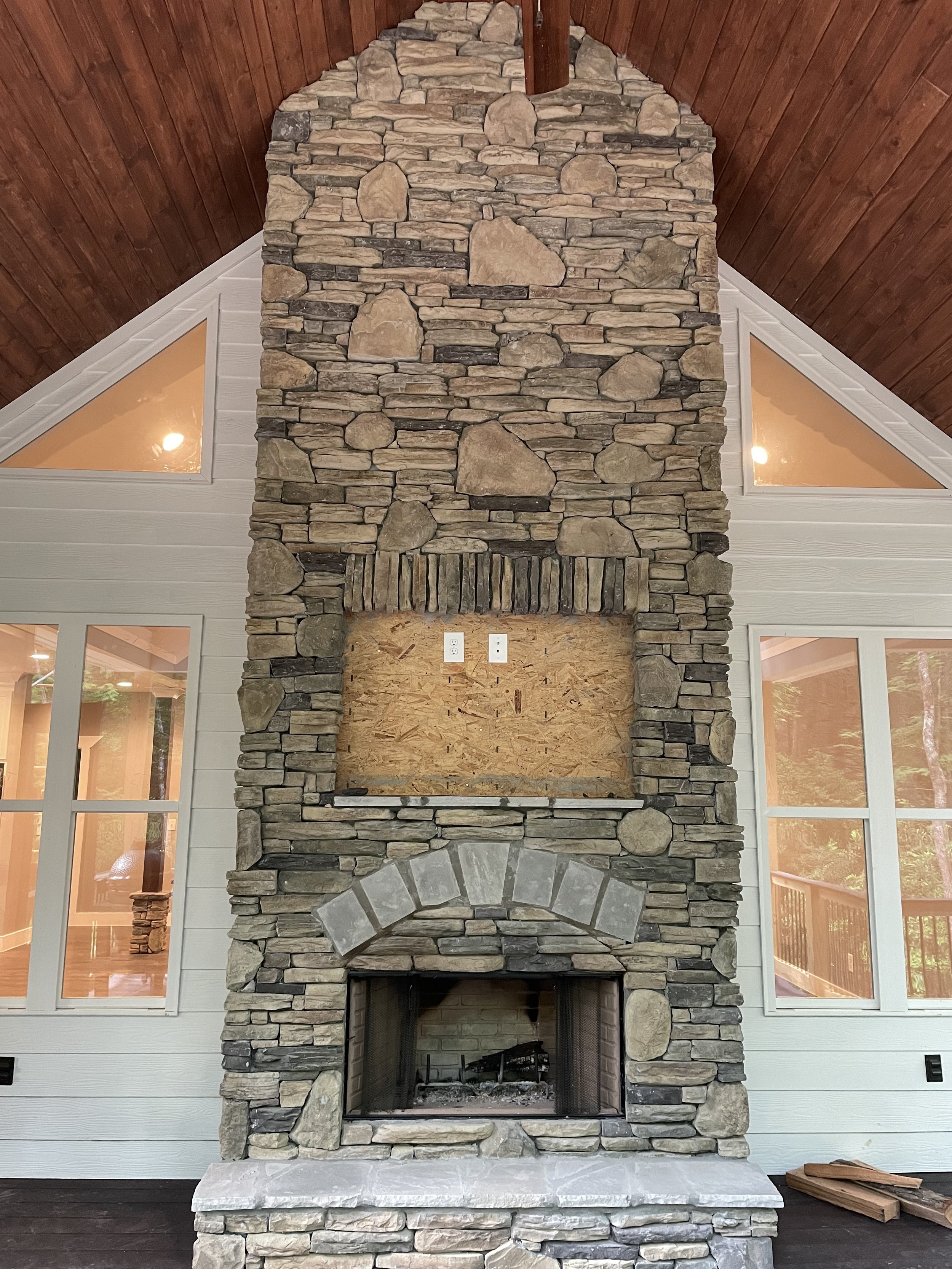 Stone fireplace with an empty wood-burning firebox, a wooden mantle, and a stenciled inset area for a TV or decoration, located between windows on a white wall, high vaulted ceiling with wood paneling.