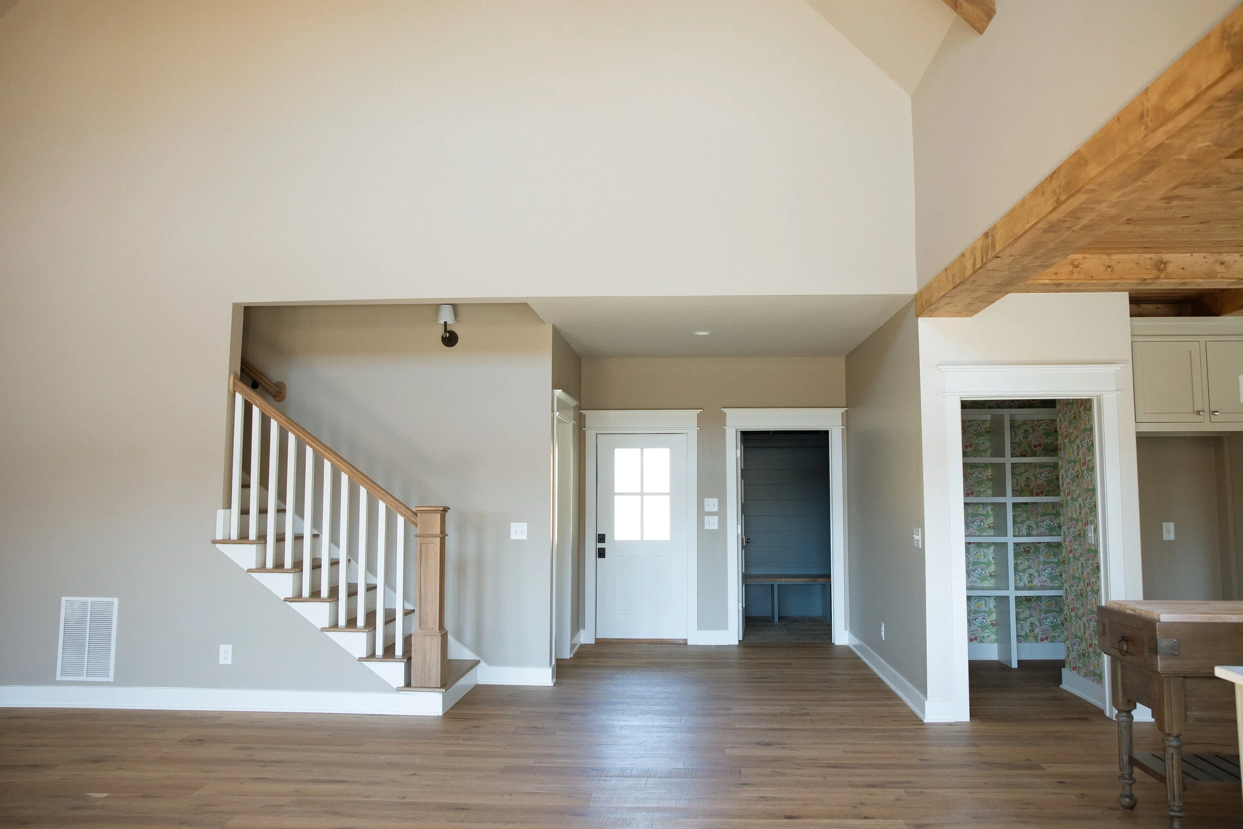 Empty living room with wooden floors, staircase on the left, and a front door with a window in the center background. A doorway with floral wallpaper interior is on the right, and a wooden beam is on the ceiling to the right.