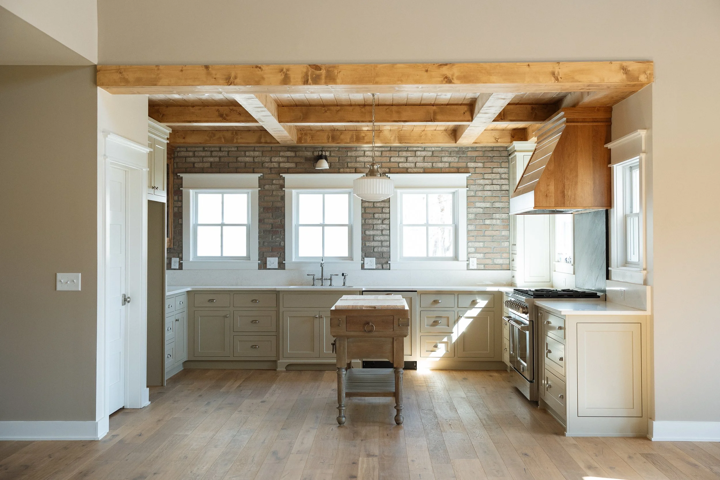 Bright kitchen with cream cabinets, brick wall, wooden ceiling beams, three windows, central island, stove, and pendant light.