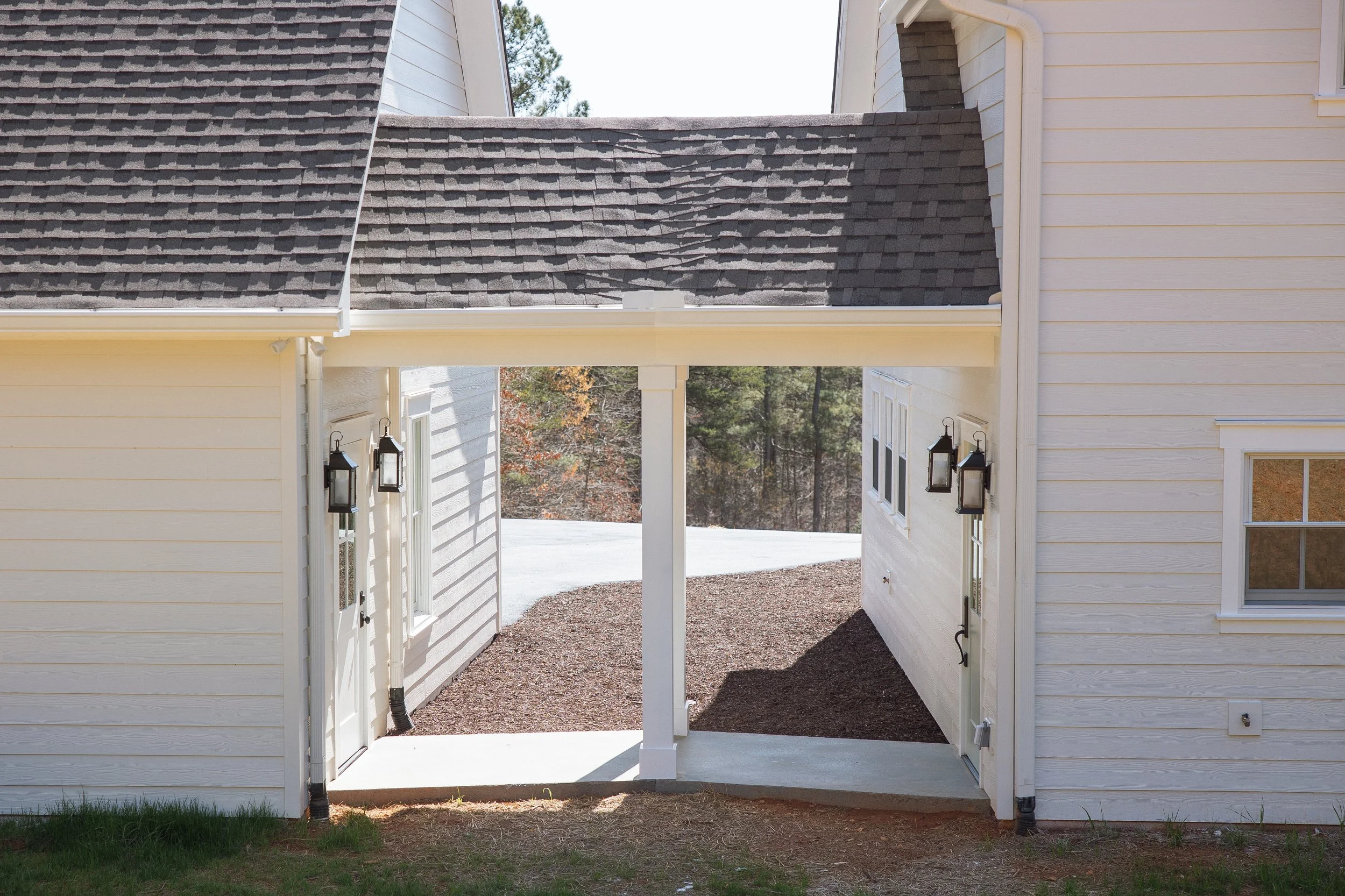 Outside view of a white house with siding, showing a covered walkway and two black lantern-style light fixtures on either side of the doorway, leading to a wooded backyard.