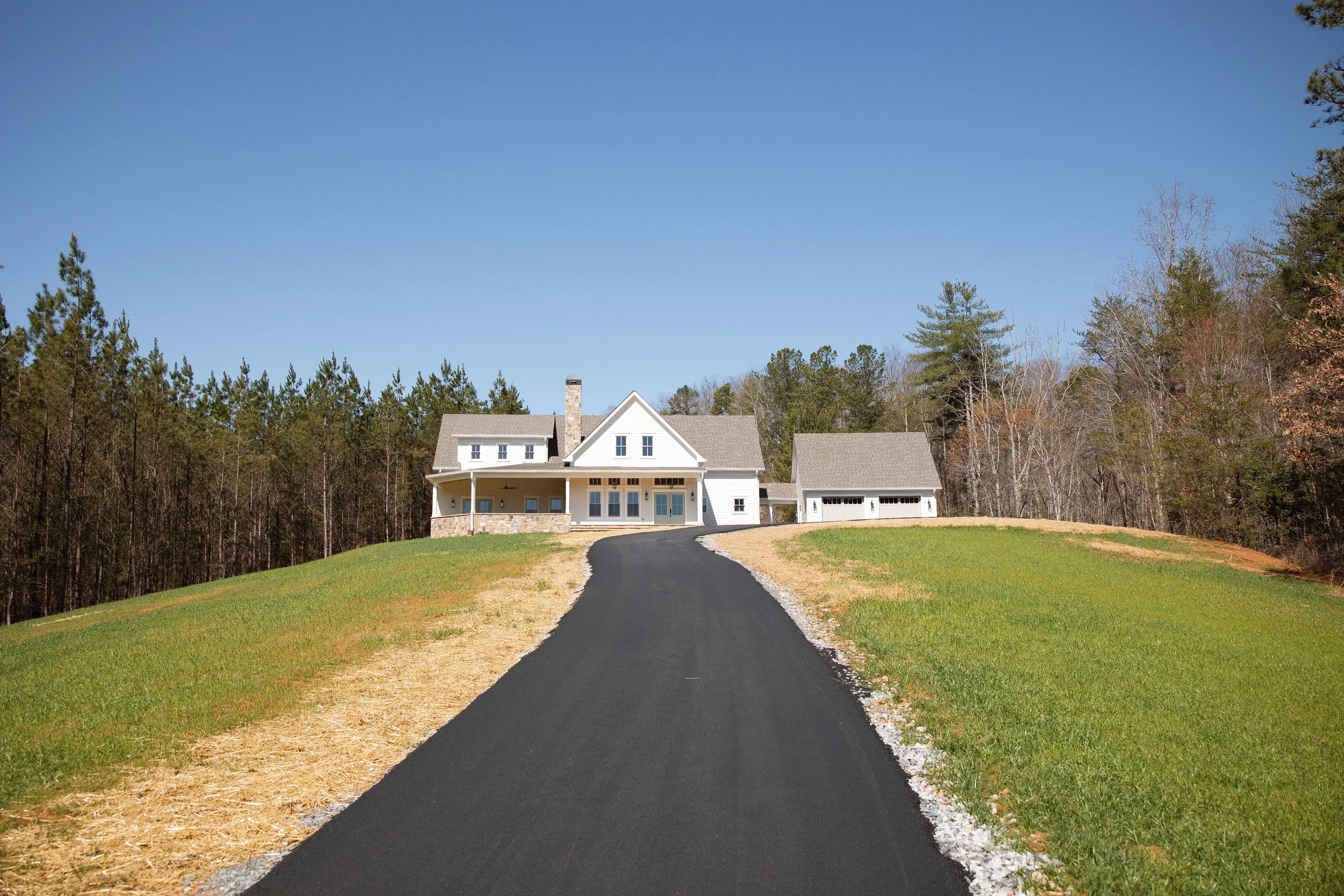 A newly paved black asphalt driveway leading up to a white house with a front porch, set on a grassy hill with a forest in the background and a clear blue sky.