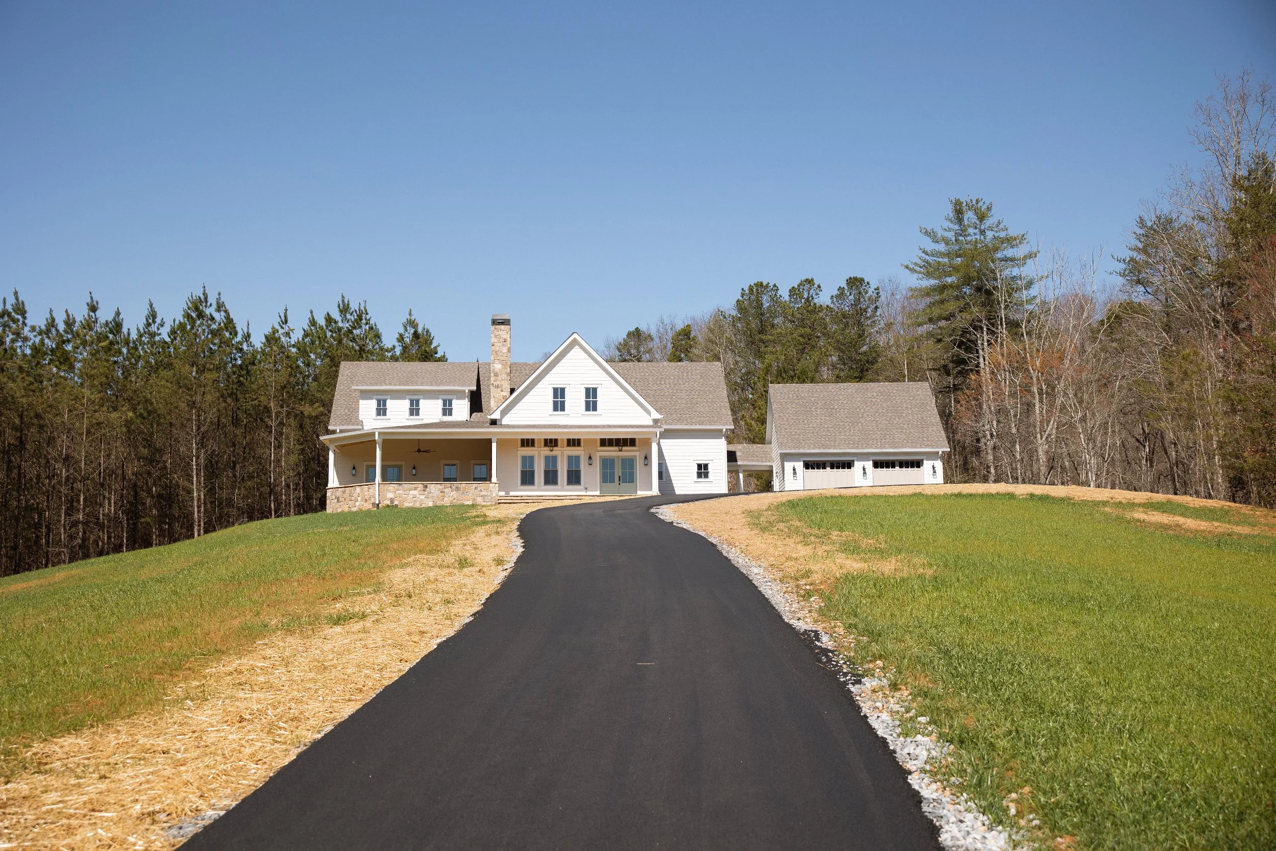 A white house with a gray roof and attached garage, situated on a grassy hill with a black asphalt driveway leading up to it, surrounded by trees and a clear blue sky.