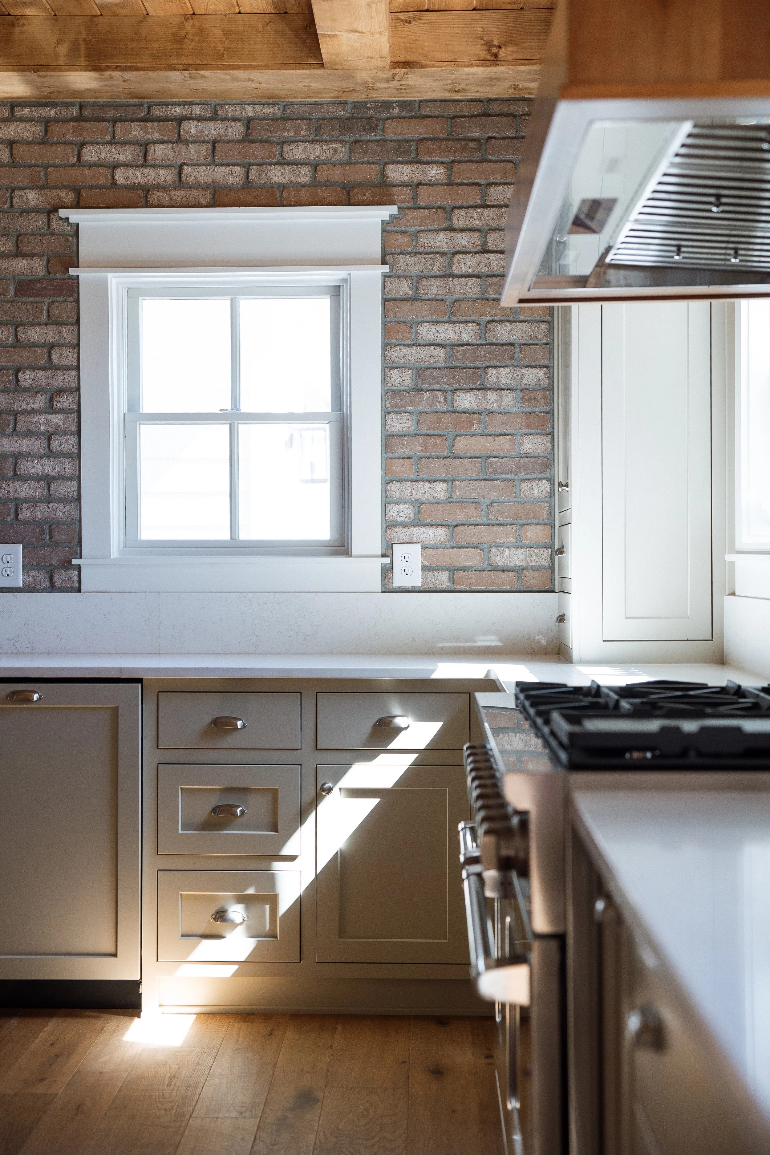A kitchen with a brick wall, a window with white trim, beige cabinets, a white countertop, and sunlight casting shadows on the floor and cabinets.