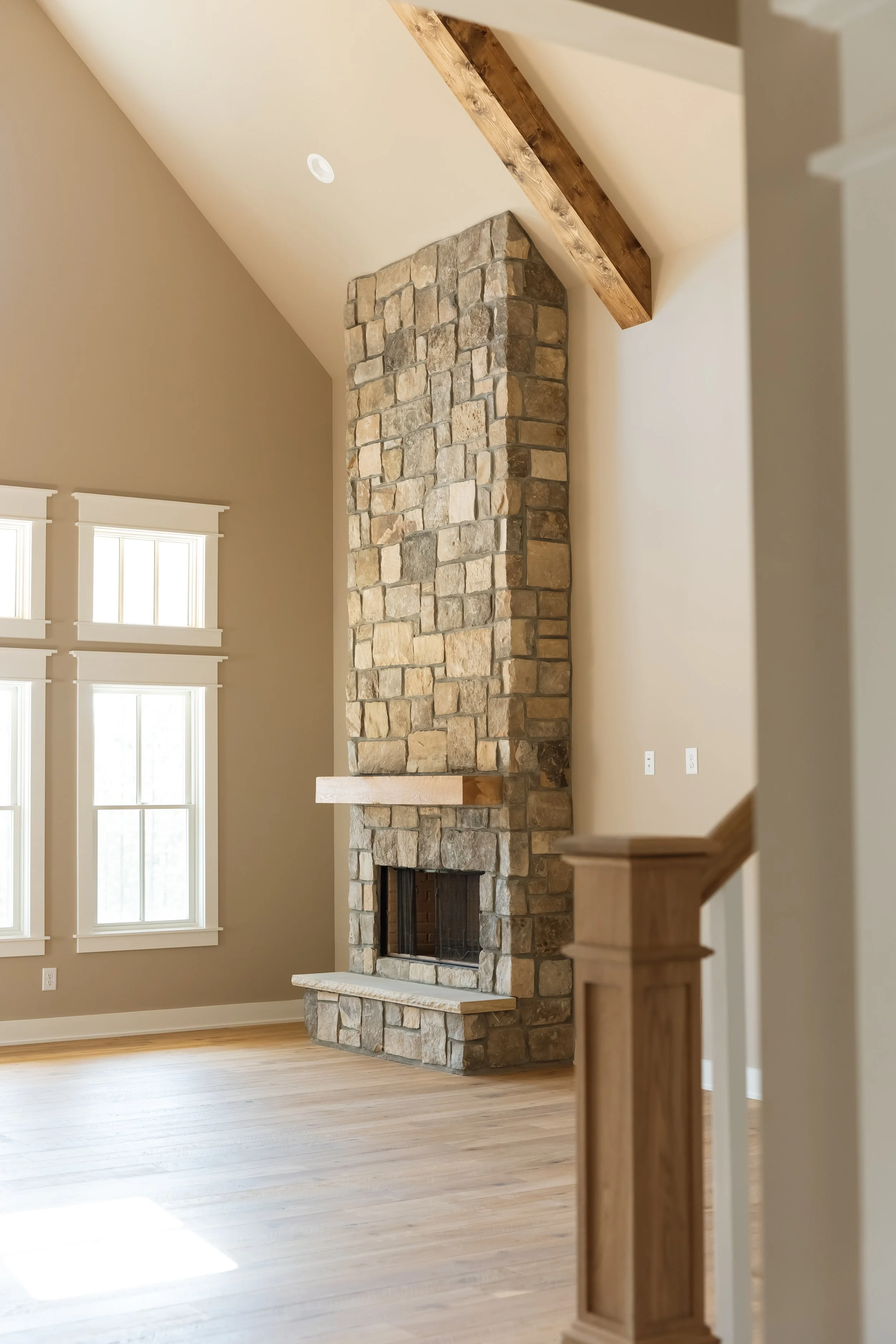 Living room with a tall stone fireplace, beige walls, three windows with white trim, hardwood floor, and a wooden beam on the ceiling.