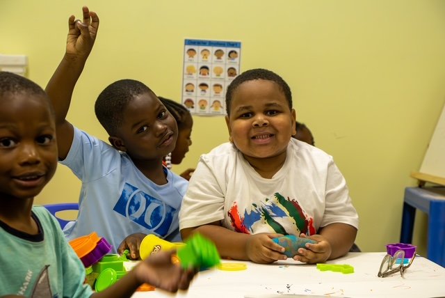 Three young children sitting at a table in a classroom, with one child raising his hand, the middle child making a grimace, and the third looking at the camera. There are toys and classroom posters in the background.