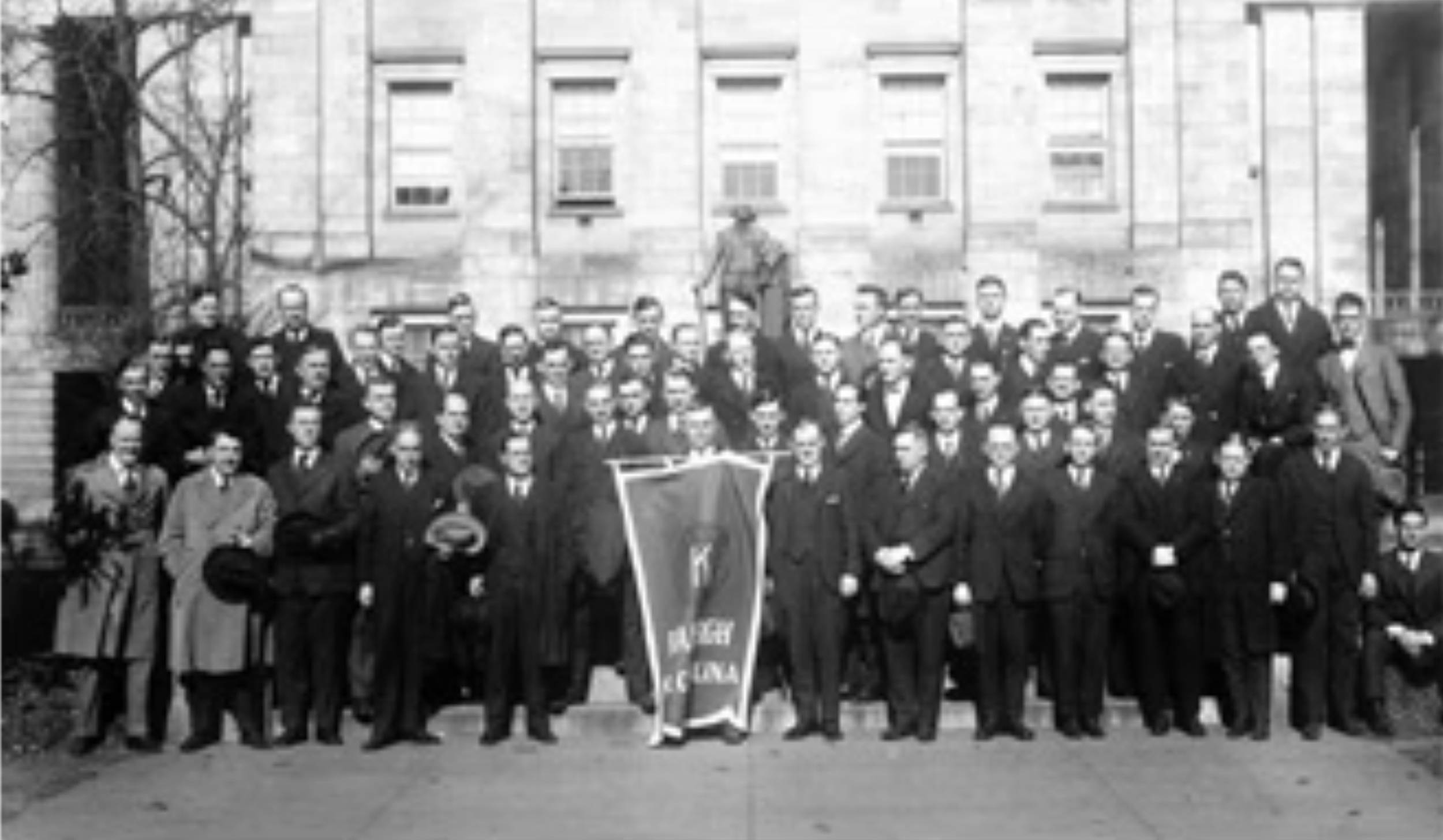 A large group of men dressed in dark suits posing in front of a building, with one man standing on a ladder and another holding a banner, during what appears to be a historical event or gathering.