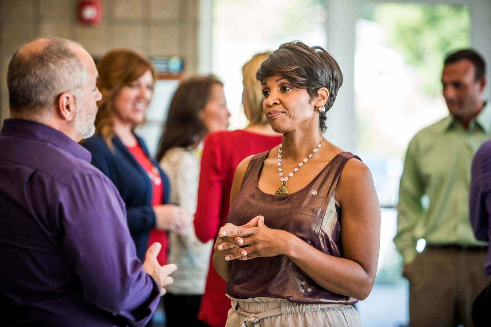 A woman in a sleeveless brown top, wearing a pearl necklace, engaged in conversation with an older man in a purple shirt, at a social gathering indoors with other people in the background.