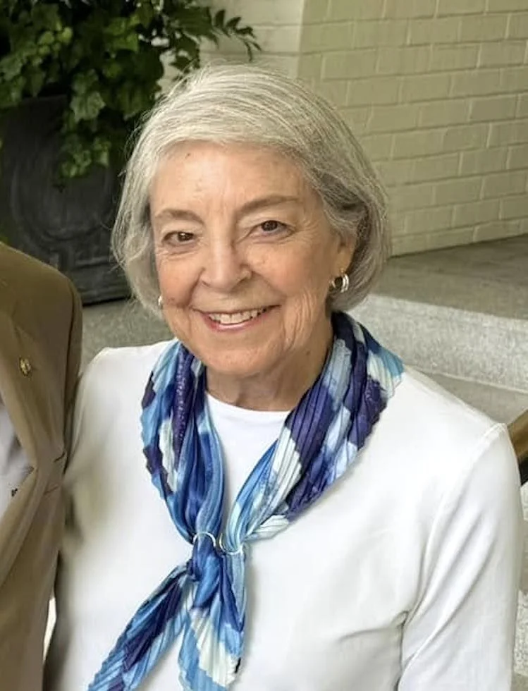 Older woman with short gray hair, wearing a white top, a blue patterned scarf, and hoop earrings, smiling at the camera, in front of a brick wall and greenery.