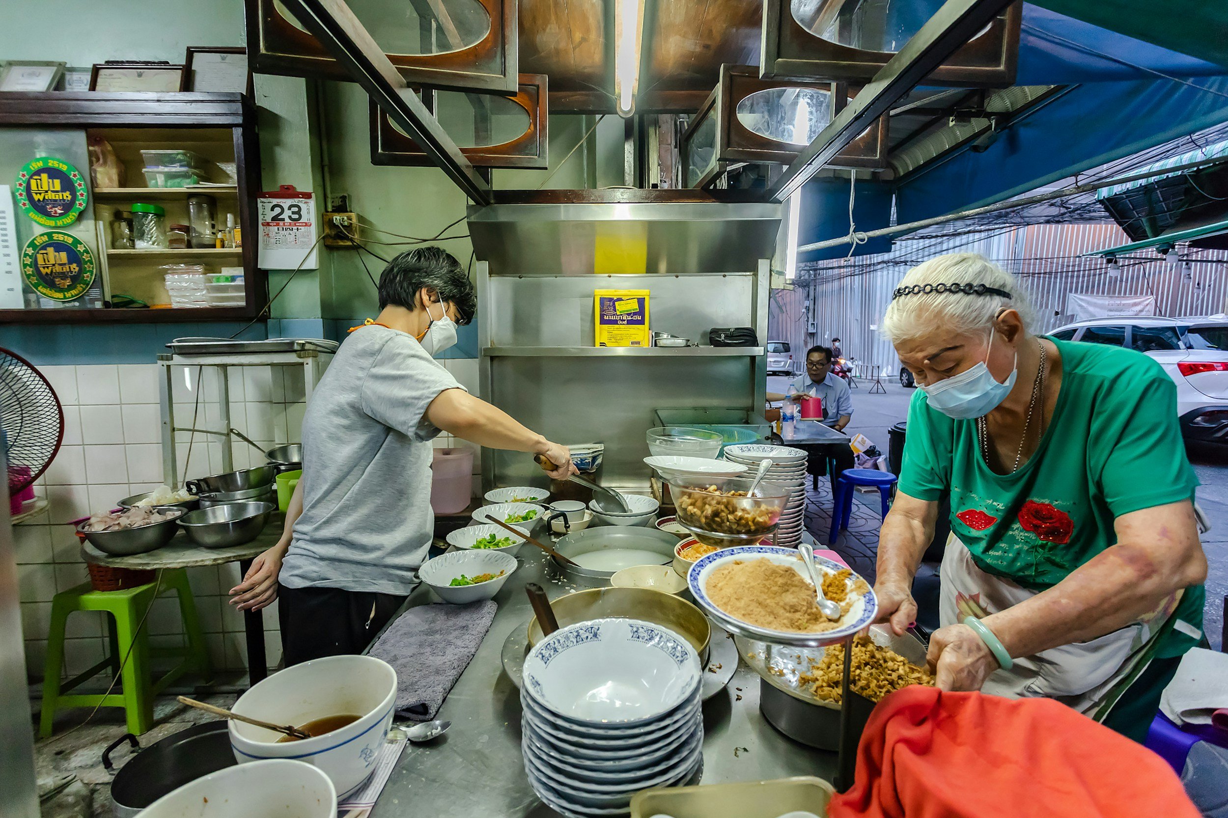 Two women preparing food at an outdoor street food stall, both wearing face masks. The woman on the right has short white hair, a headband, and is wearing a teal shirt with red lips printed on it. The woman on the left has short dark hair, glasses, and is wearing a gray shirt. There are various bowls and ingredients on the counter, and parking lots and cars are visible in the background.