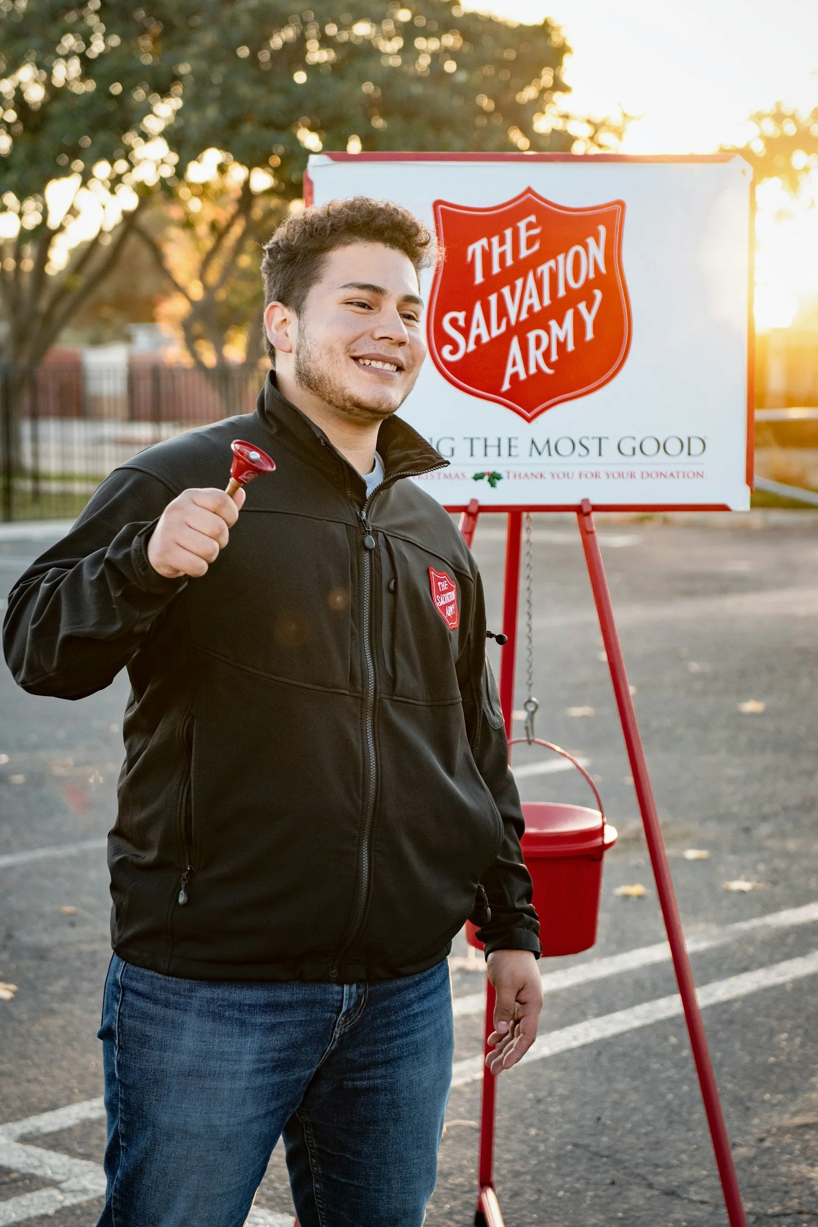 A young man smiling and holding a small red bell in front of a Salvation Army donation stand outdoors during sunset.