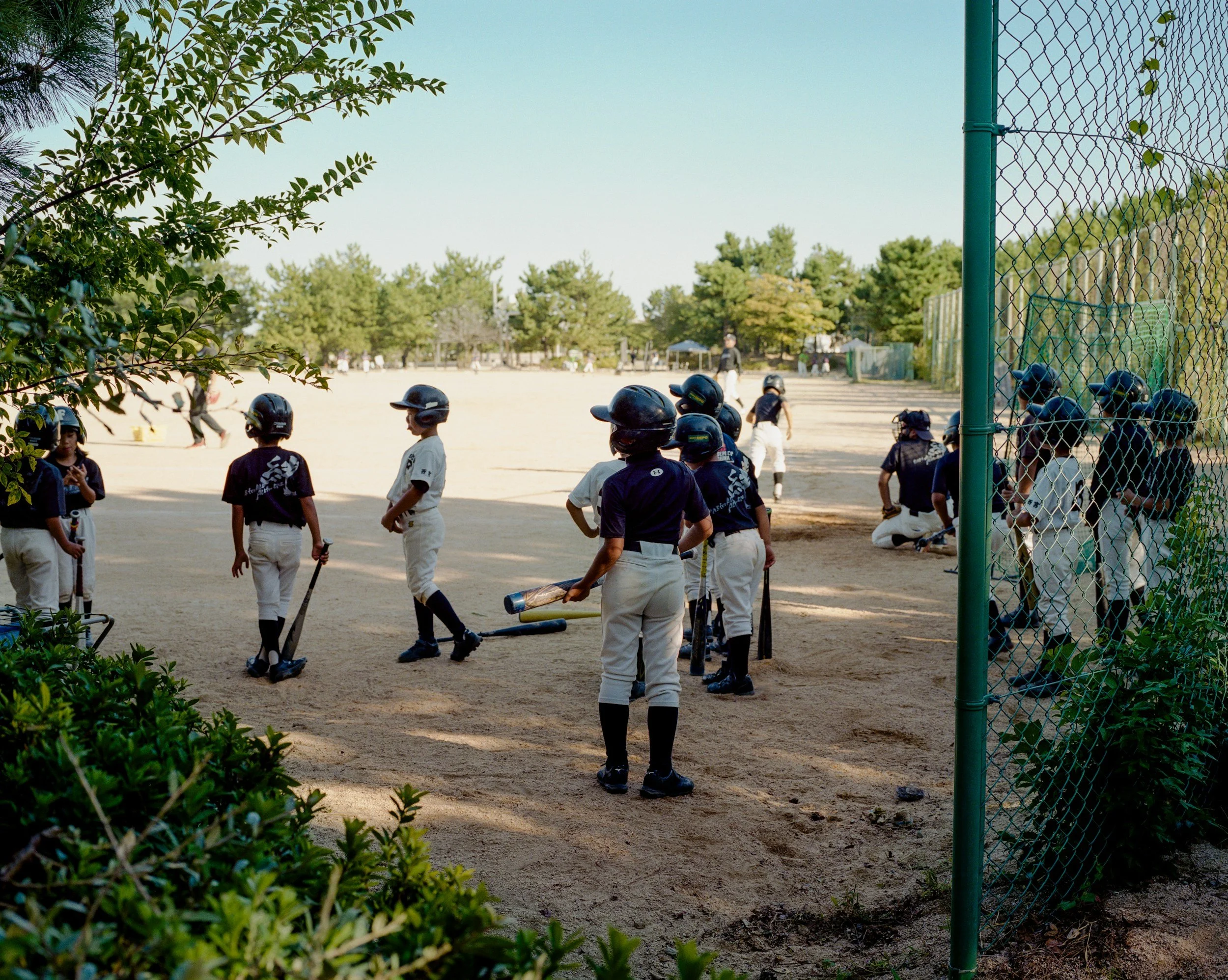 Boys in baseball uniforms with helmets on, gathered on a dirt baseball field, some holding bats, others sitting or standing, surrounded by trees and a chain-link fence.