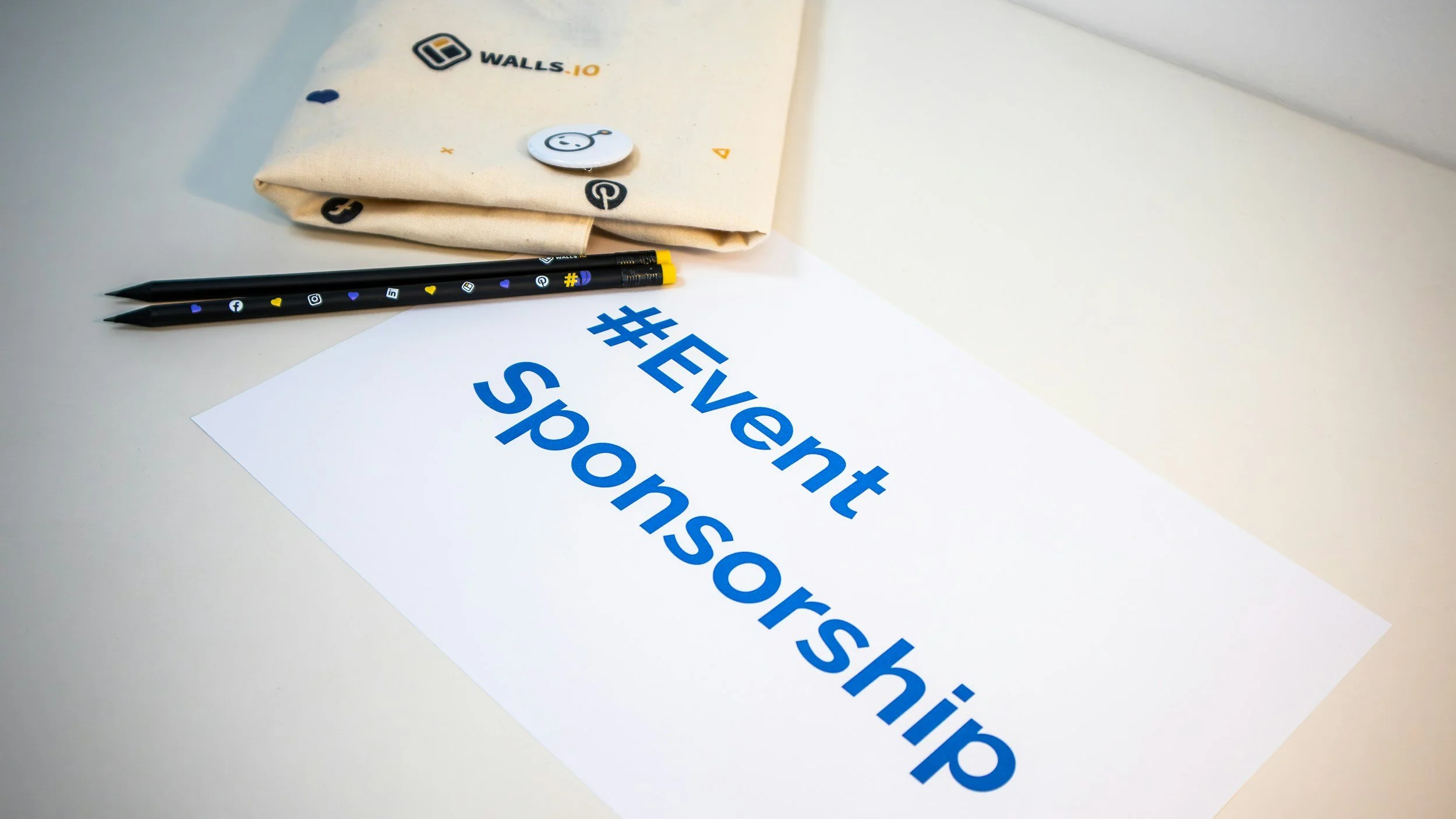 A white sheet of paper with blue text reading '#Event Sponsorship' on a white surface, two black pens with social media icons, a beige cloth bag with logo and small button with smiley face, and a napkin with logo.