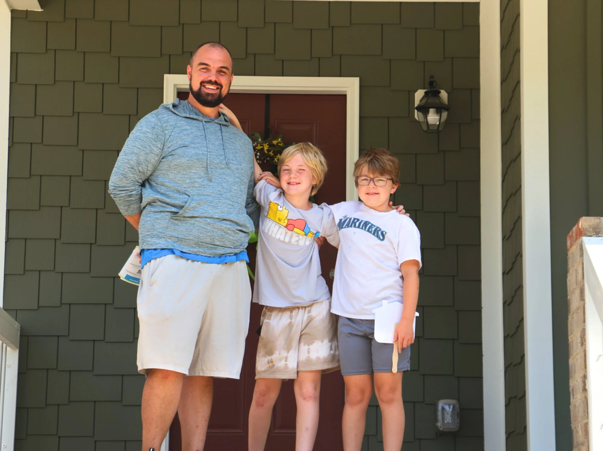 A smiling man with a beard stands on the porch with two children, all with arms around each other, in front of a green house with a red door and exterior light fixture.