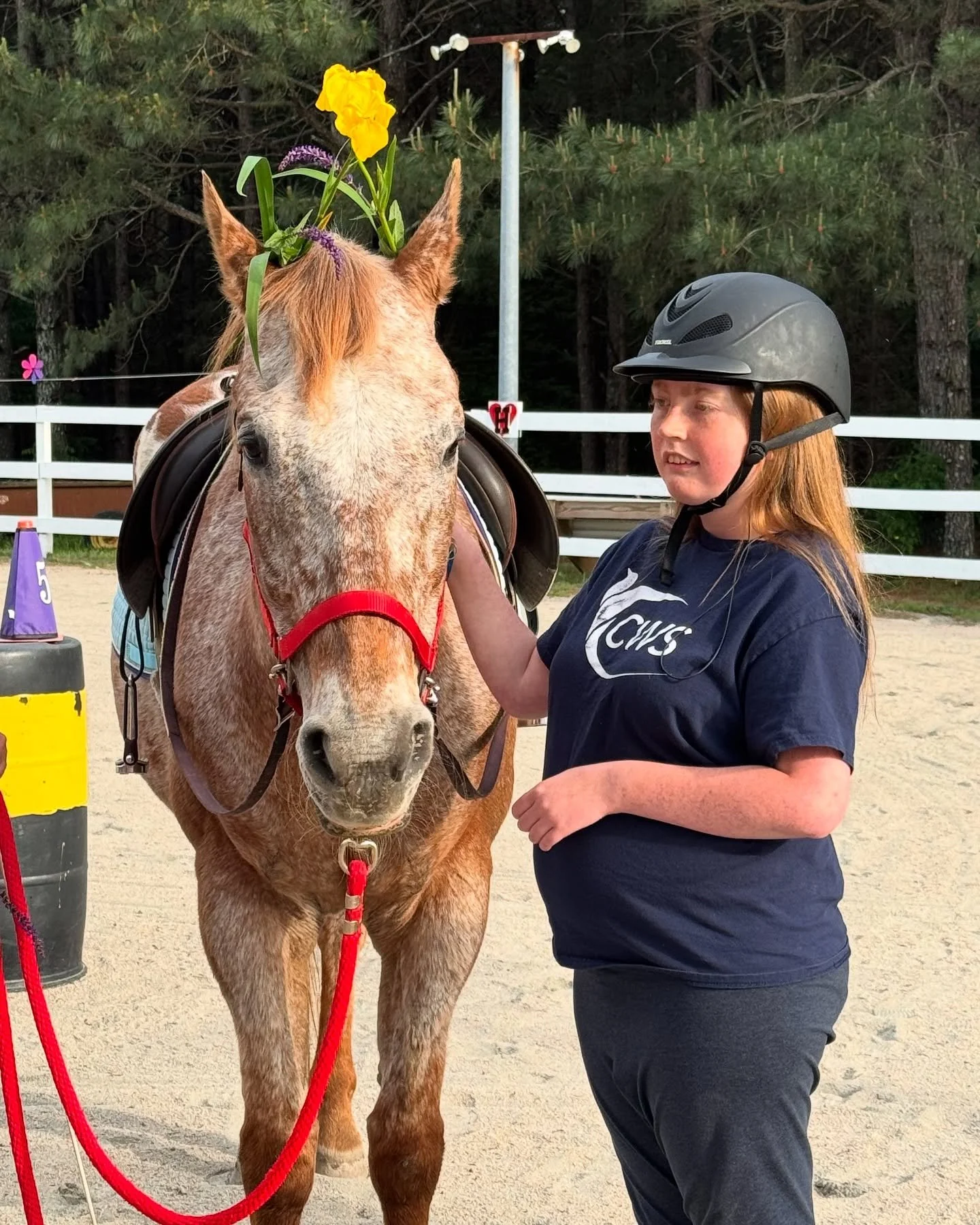 A girl wearing a helmet and a navy blue shirt stands next to a light-colored horse with a flower crown on its head, in an outdoor riding arena.