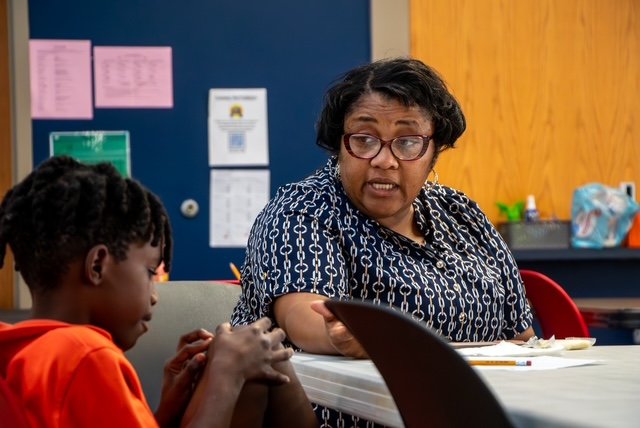 A woman with glasses and short black hair talking to a young girl with braided hair in a classroom.