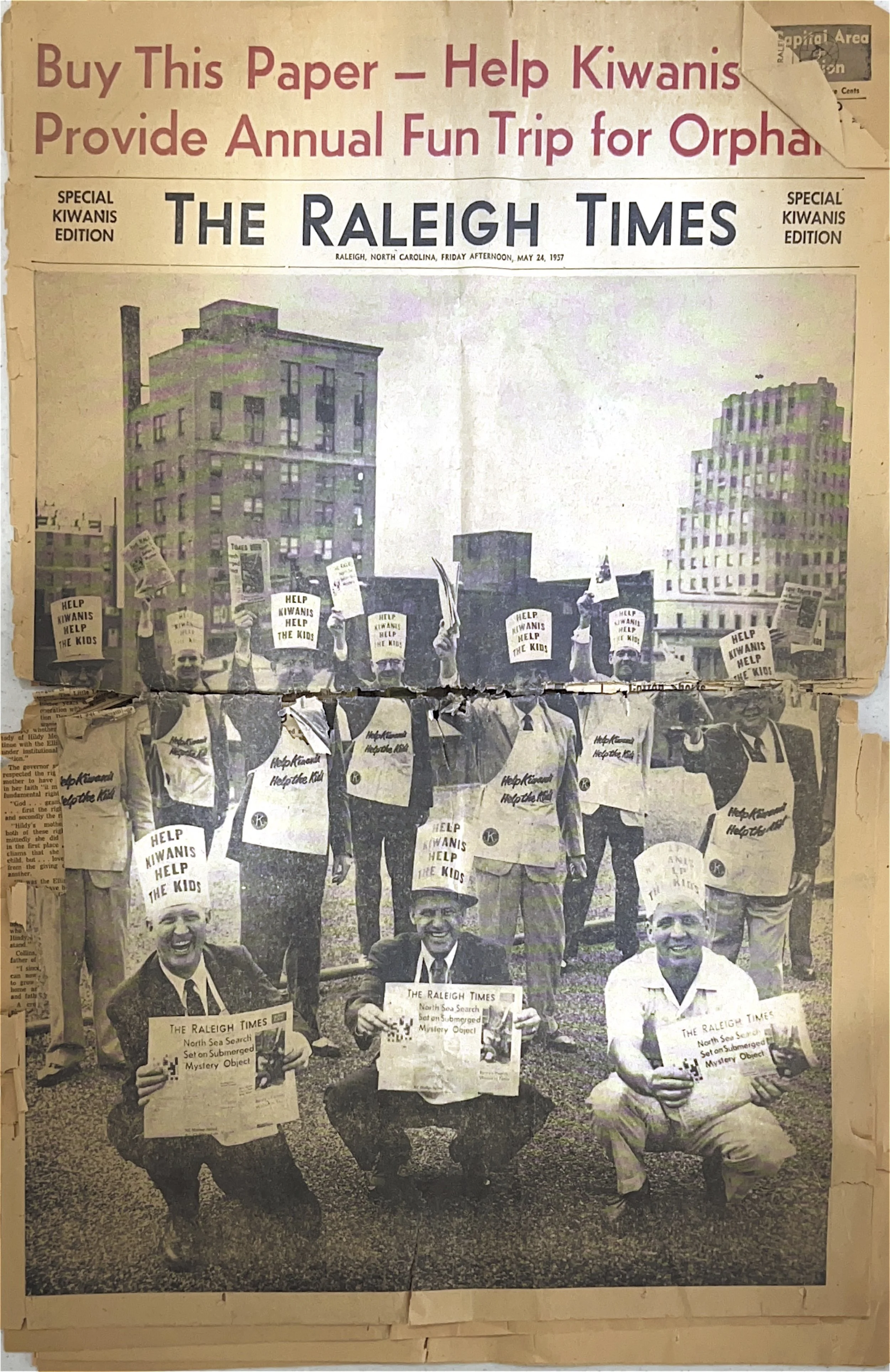 A black and white newspaper front page with a photo of to a group of people holding signs and newspapers, protesting for help for Kiwanis and orphans. The headline indicates it is from The Raleigh Times, dated May 24, 1957.