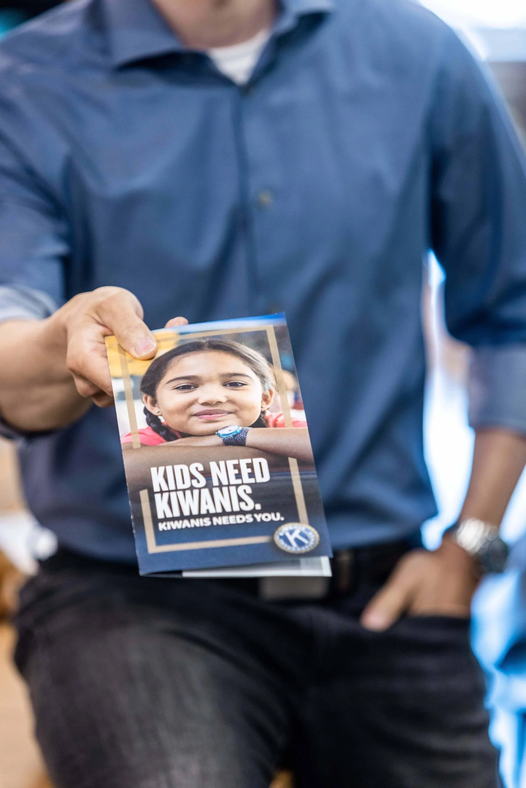 A person holds a flyer with a photo of a girl and text that reads 'Kids need Kiwanis. Kiwanis needs you.'
