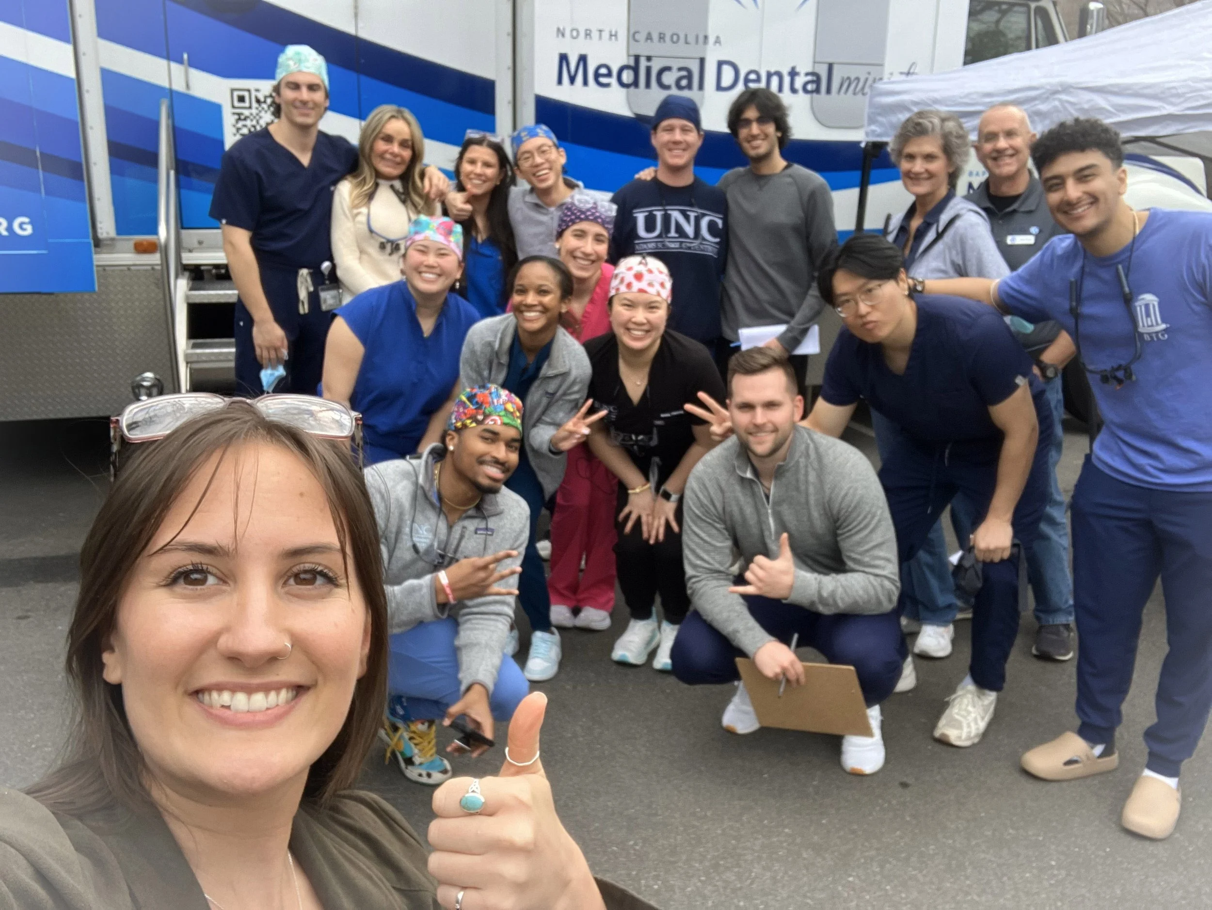 Group of medical professionals and volunteers posing for a photo outdoors in front of a mobile dental clinic truck, smiling and making gestures toward the camera.
