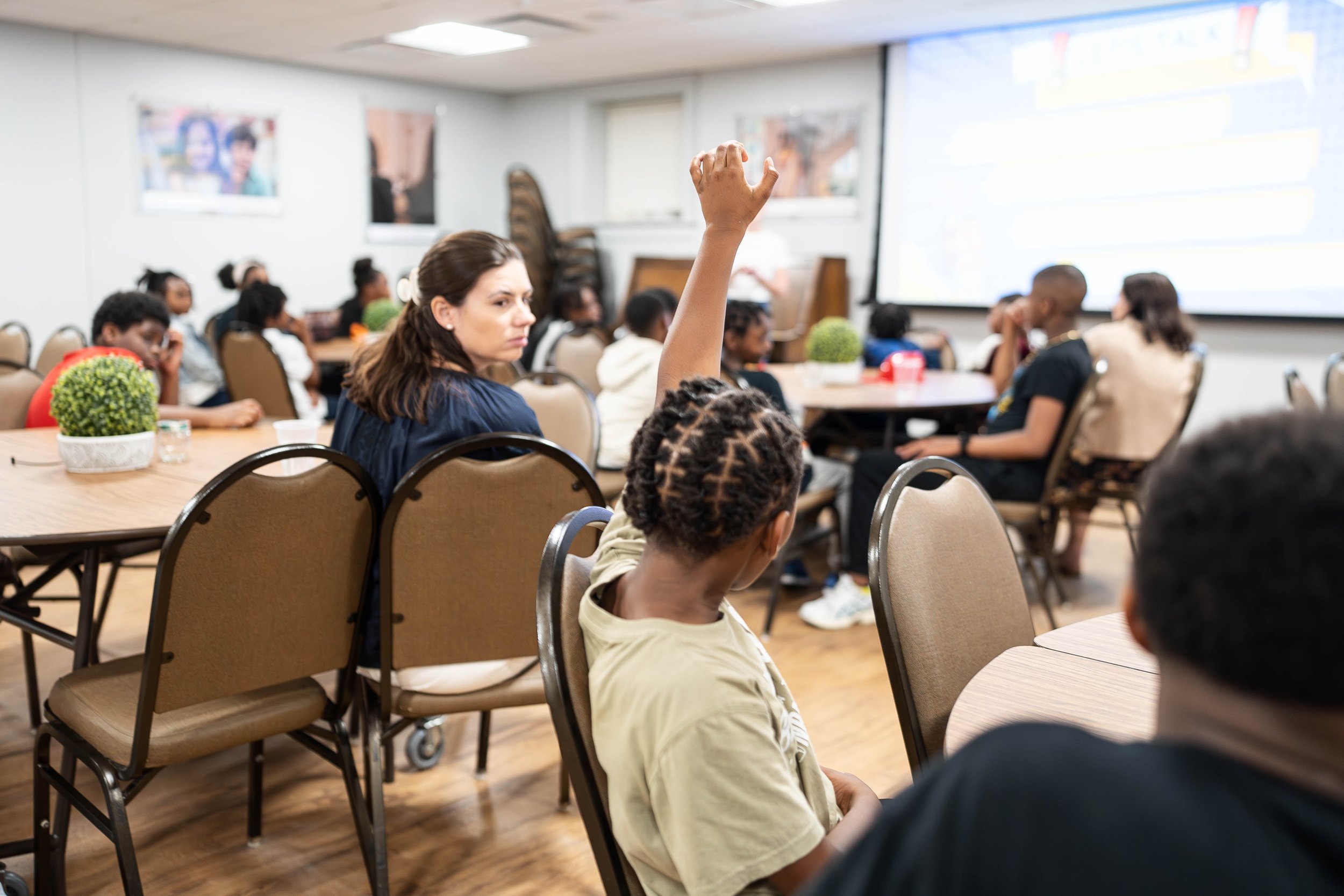 A young girl with braided hair raises her hand in a classroom setting filled with students and a teacher sitting at tables, some students are seen listening attentively and a woman looks thoughtfully towards the front.