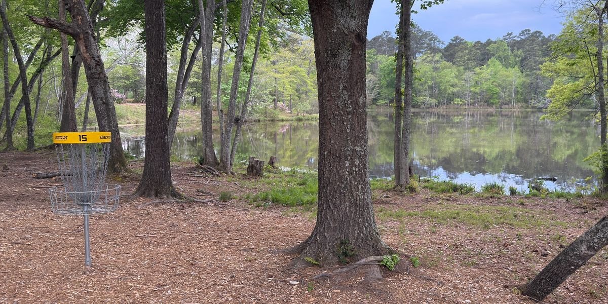A disc golf basket with yellow top and numbered 15, situated among trees near a calm lake in a wooded area.
