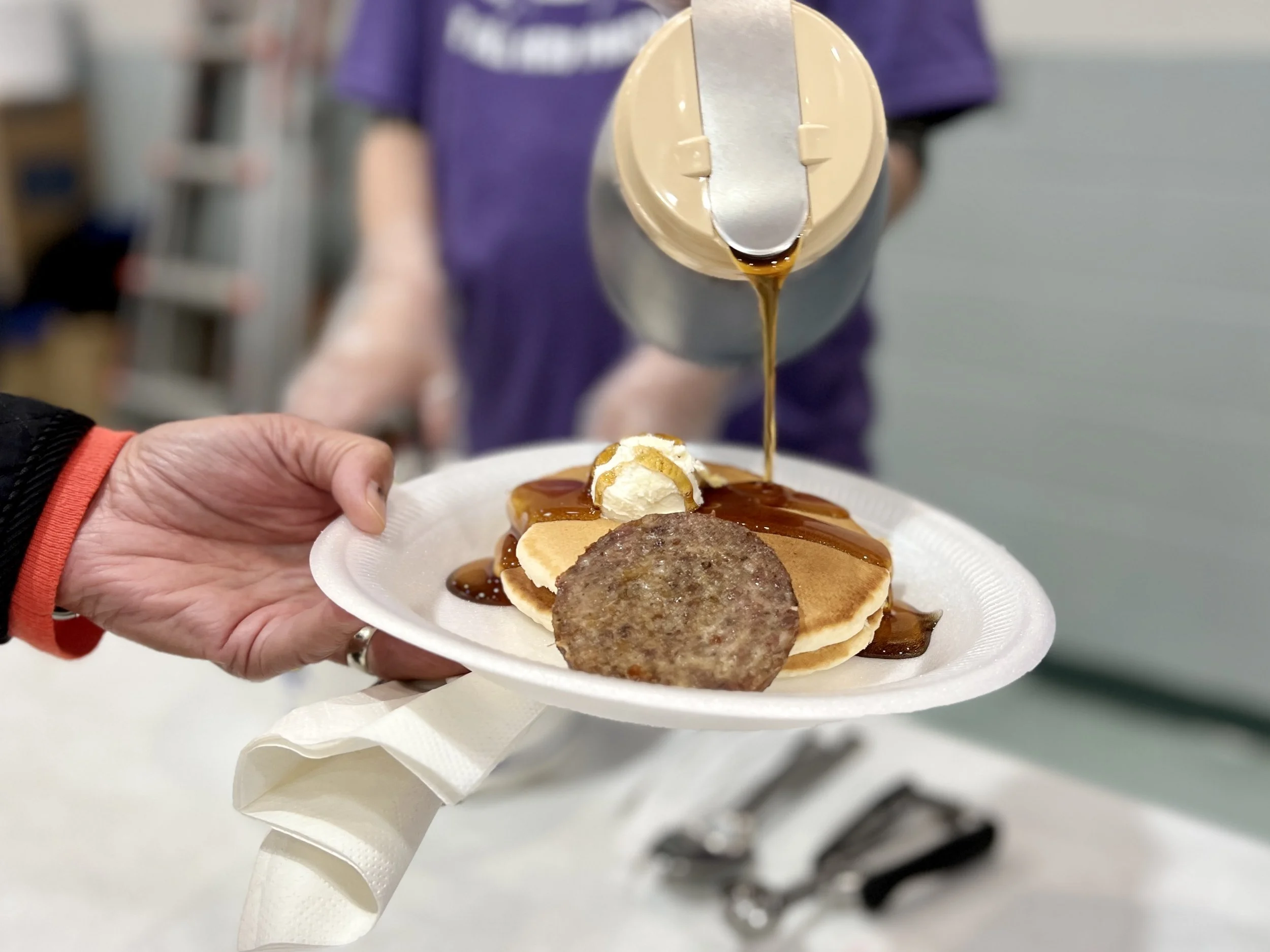 Pouring syrup over stacked pancakes topped with butter and sausage patty on a paper plate held by a person, with another person in the background.